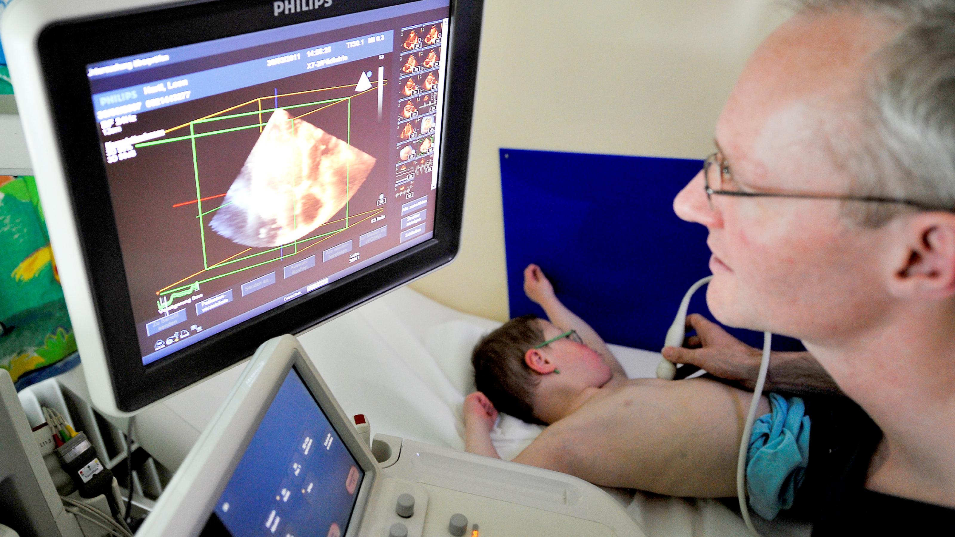 Ein Mann betrachtet auf einem Monitor das Ultraschallbild eines Herzen. Den Fühler hält er einem Jungen auf die Brust, der neben ihm liegt.