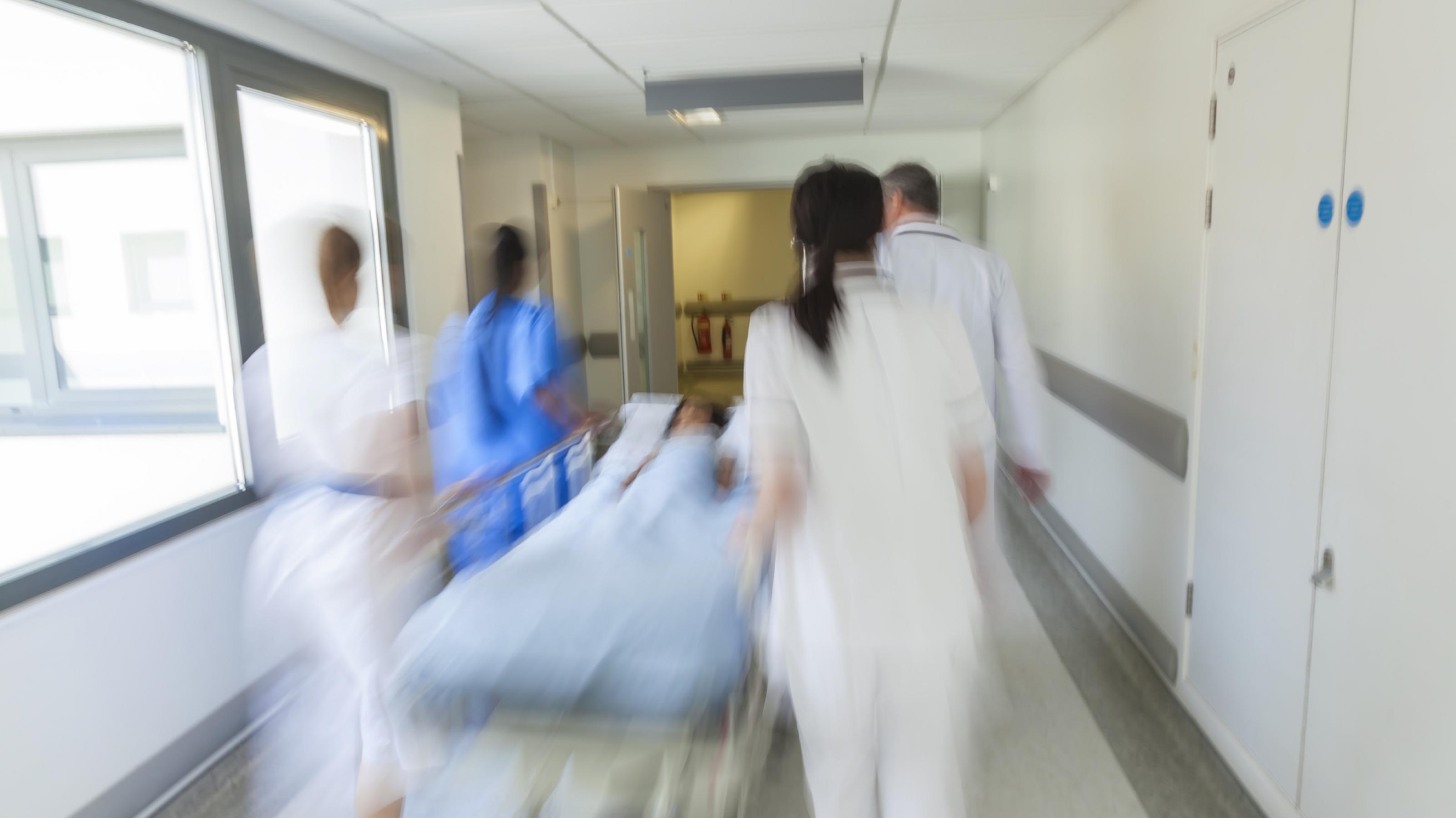 Das Bild zeigt medizinisches Personal, das einen Patienten in einem Bett durch die Klinik schiebt. Das Symbolfoto ist unscharf und soll Eile darstellen.