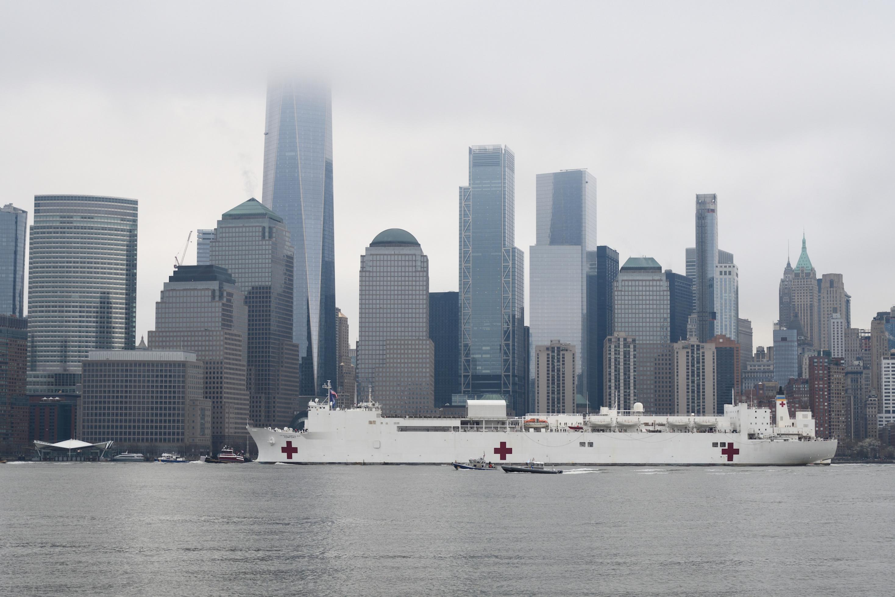 Das Bild zeigt ein großes weißes Schiff mit Rotem Kreuz vor der Kulisse von Manhattan.