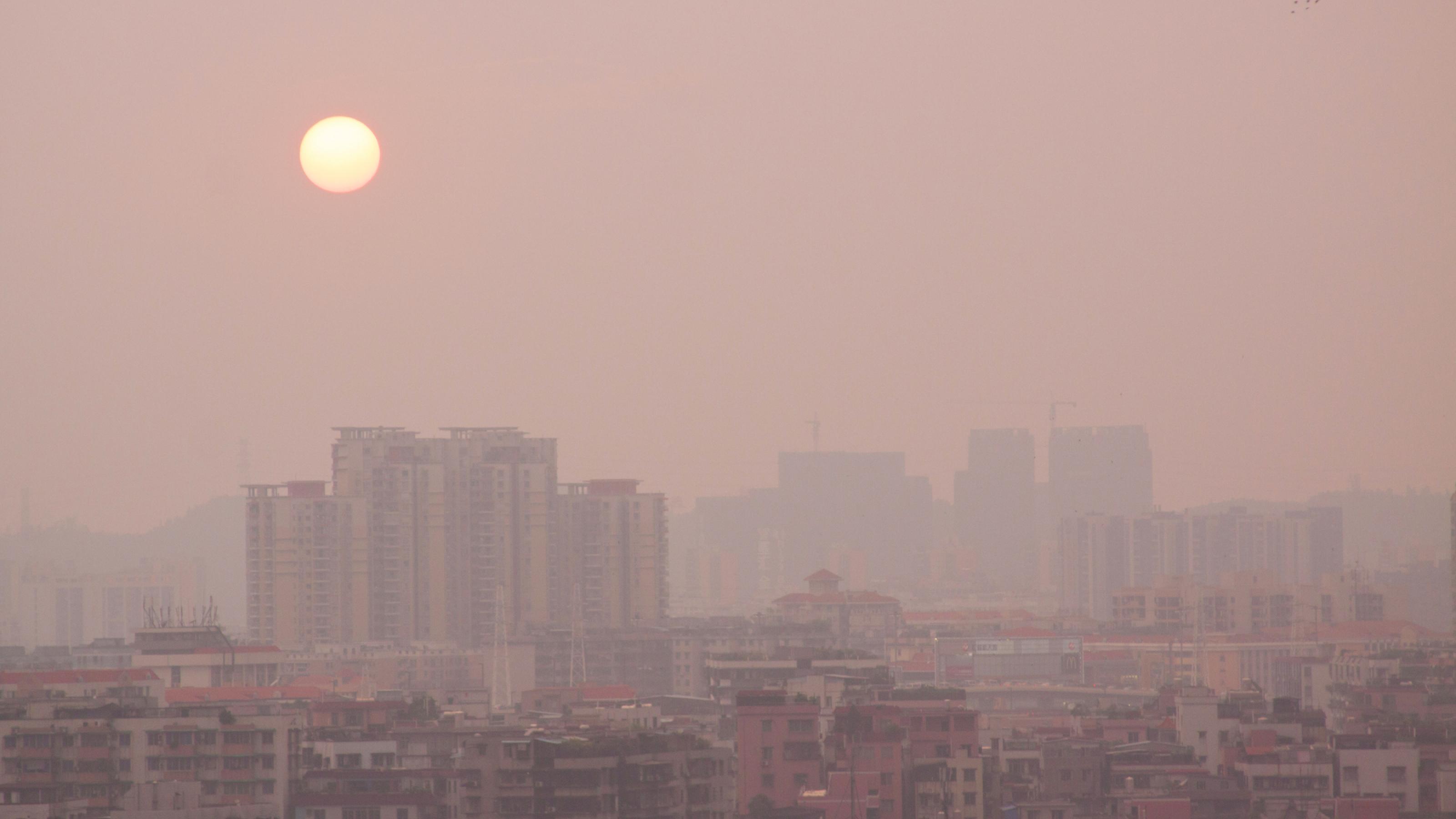 Das Bild zeigt die Silhouette der chinesischen Stadt Guangzhou mit Hochhäusern. Über der Stadt liegt eine Schicht an Smog, hinter der die Sonne nur blass zu sehen ist.