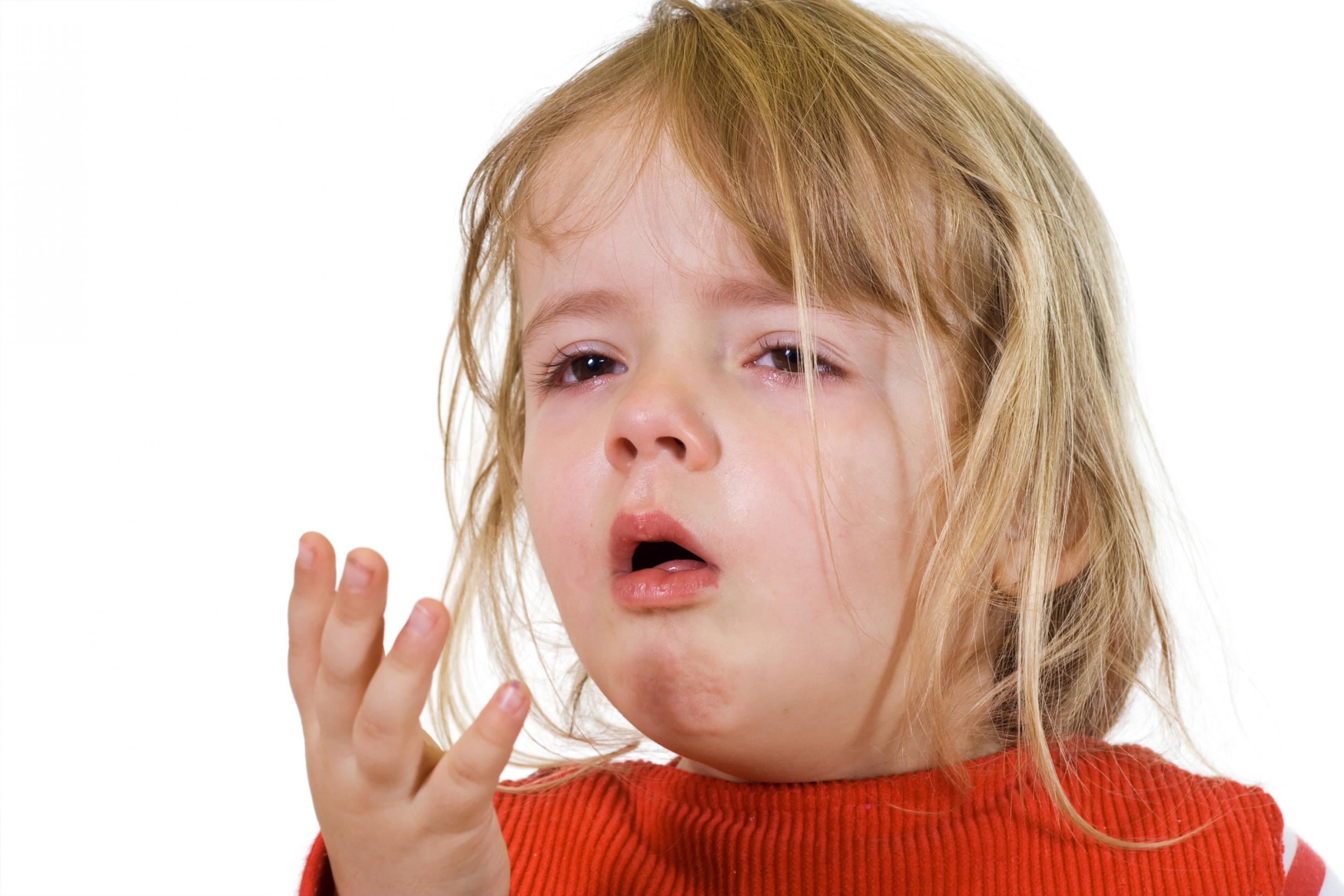 Ein etwa dreijähriges Mädchen mit braunen Haaren und einem roten Pulli hält sich die Hand vor den Mund, weil es husten muss.