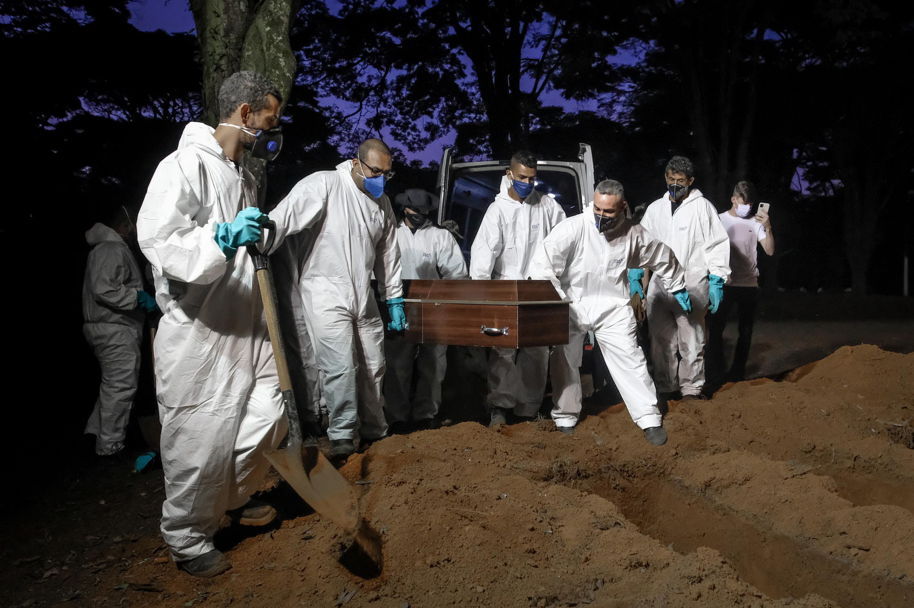 Menschen in weißer Schutzkleidung und mit Gesichtsmasken hieven bei nächtlichem Licht einen Sarg aus einem Beerdigungsfahrzeug und tragen ihn zum frisch ausgehobenen Grab.