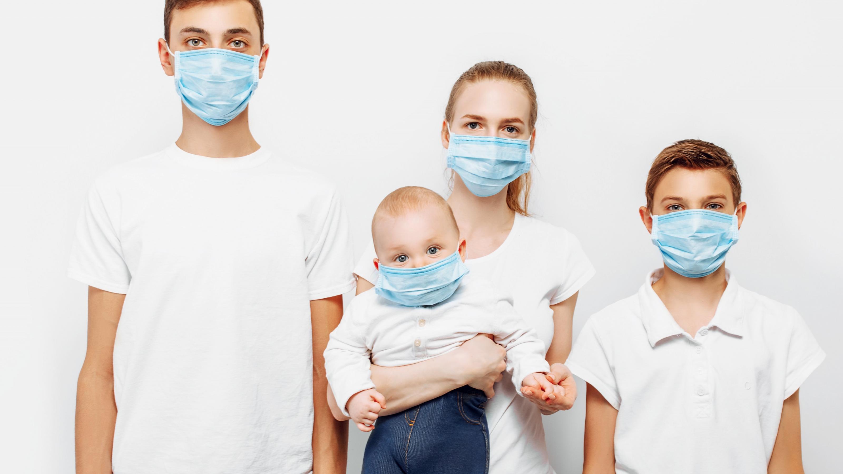 Junge Eltern mit einem Baby und einem älteren Sohn. Alle tragen eine Maske zum Schutz vor einer Corona-Infektion und schauen direkt in die Kamera.