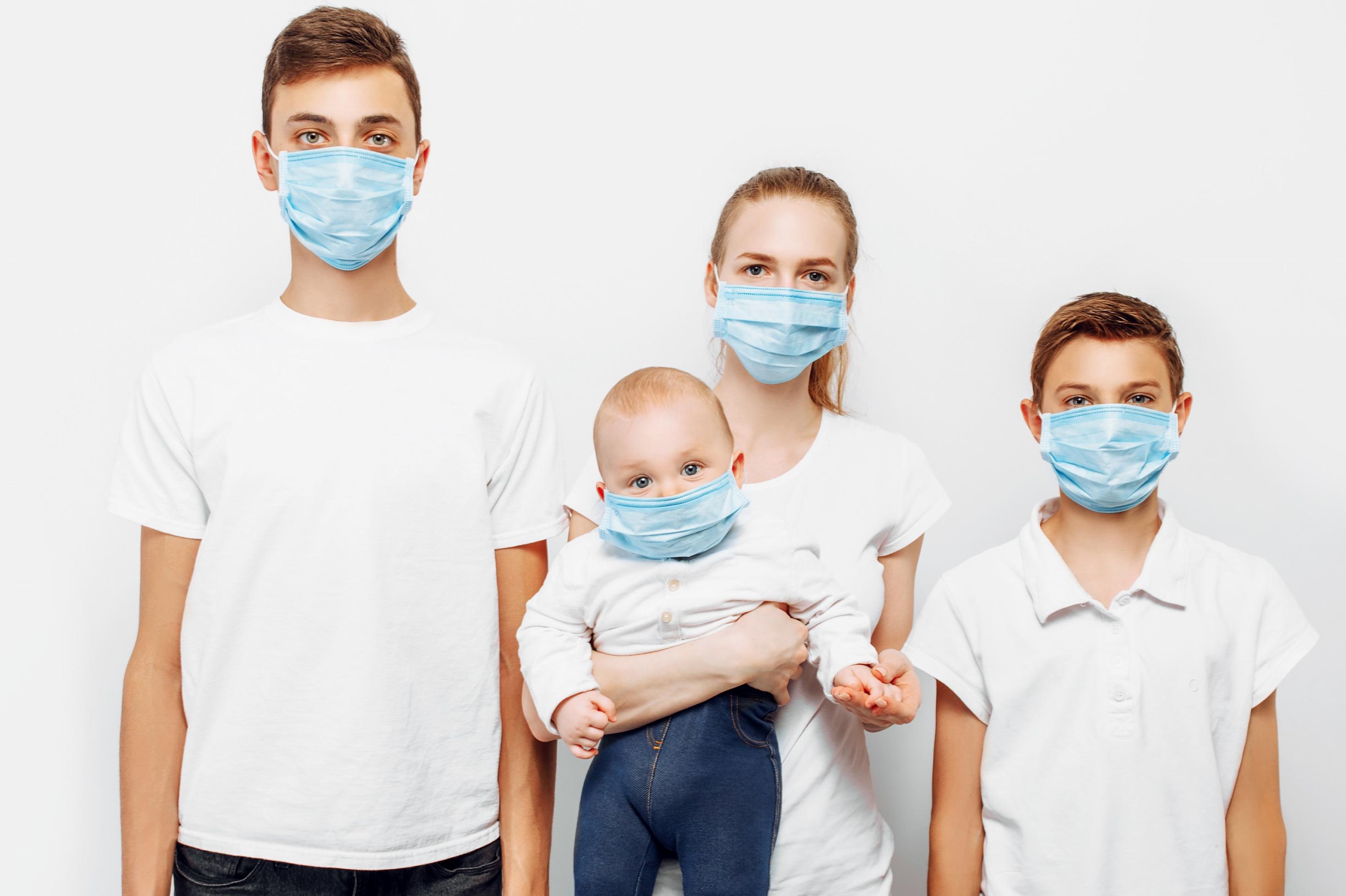 Junge Eltern mit einem Baby und einem älteren Sohn. Alle tragen eine Maske zum Schutz vor einer Corona-Infektion und schauen direkt in die Kamera.