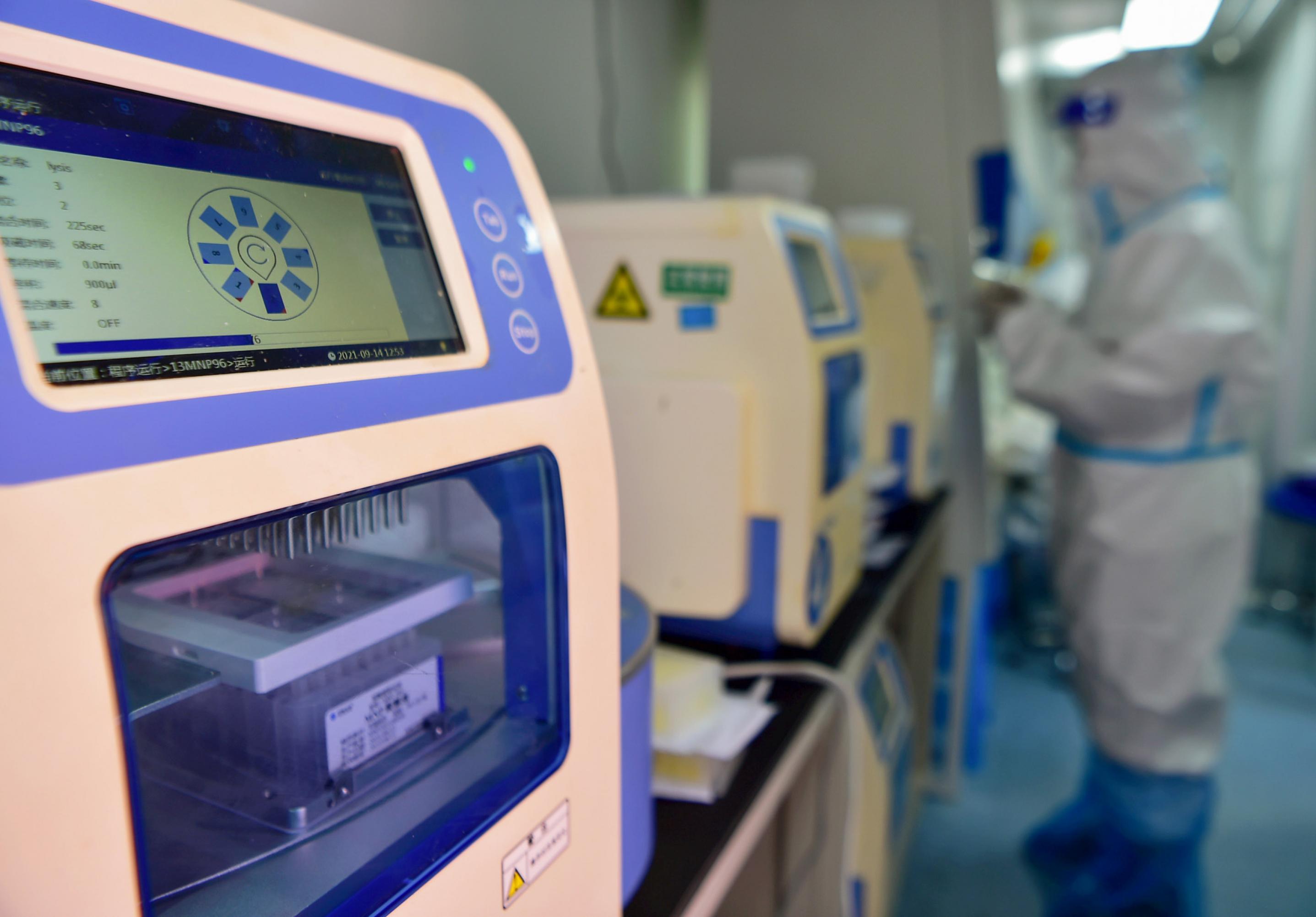 Eine beige PCR-Maschine mit großem Display. Hinter einer Scheibe sind Röhrchen zu sehen. Im Hintergrund ein Mensch, der von Kopf bis Fuß in einen Gummianzug gepackt ist.