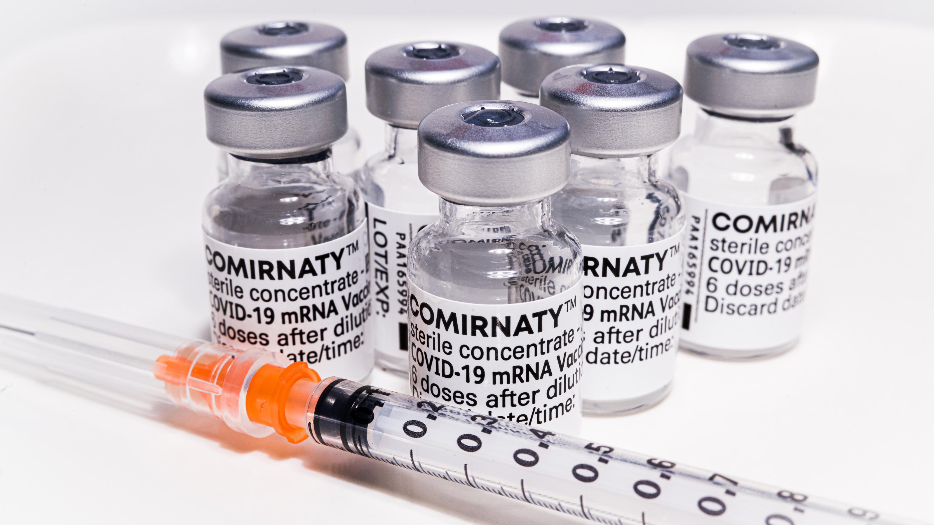 Auf dem Foto sind mehrere Ampullen mit dem mRNA-Impfstoff Corminaty abgebildet. Darunter liegt eine Impfspritze.