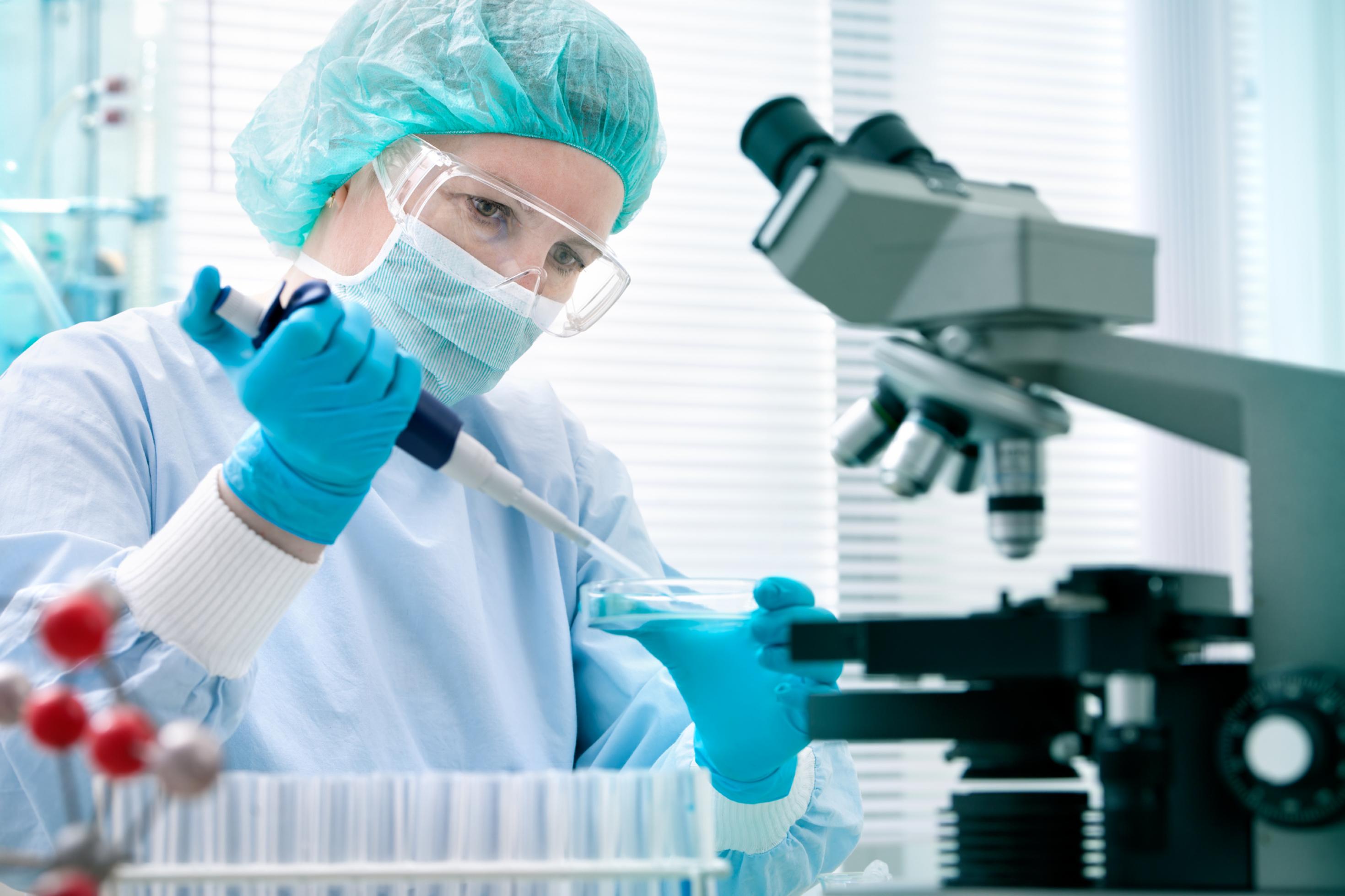 Das Symbolfoto zeigt eine Wissenschaftlerin im Labor. Sie trägt einen Mundschutz und hat eine Pipette in der Hand, mit der sie etwas in eine Petrischale träufelt. Auch ein Mikroskop ist auf dem Bild dargestellt.