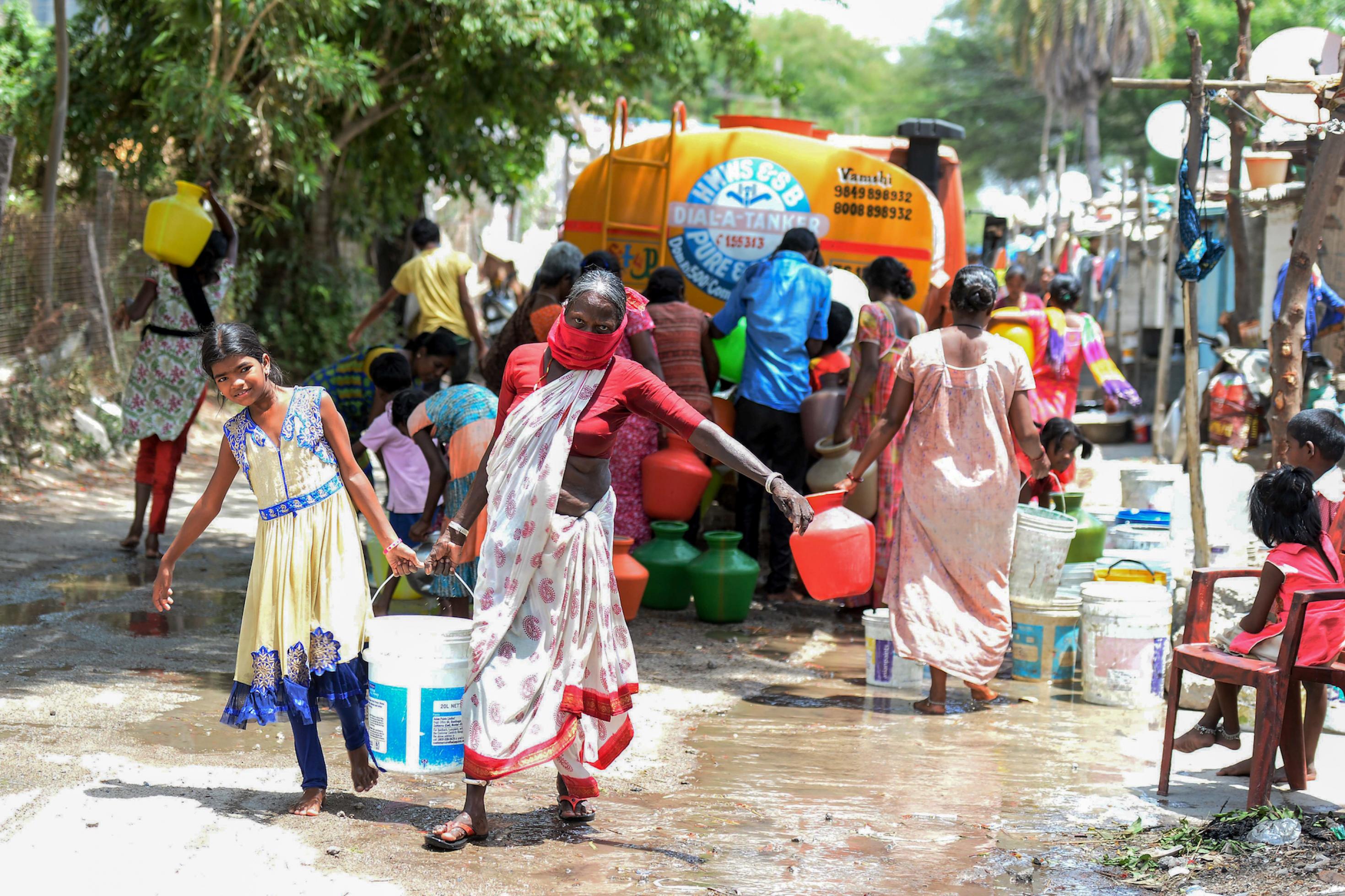 Die gezeigte Szene stammt aus dem Mai 2020. In einem Slum im indischen Secunderabad bringt ein Lastwagen kostenloses Trinkwasser. Viele Menschen stehen um den Lastwagen herum und versuchen, etwas abzubekommen. Eine Frau und ein Mädchen waren erfolgreich. Sie tragen einen großen Wasserbehälter nachhause.
