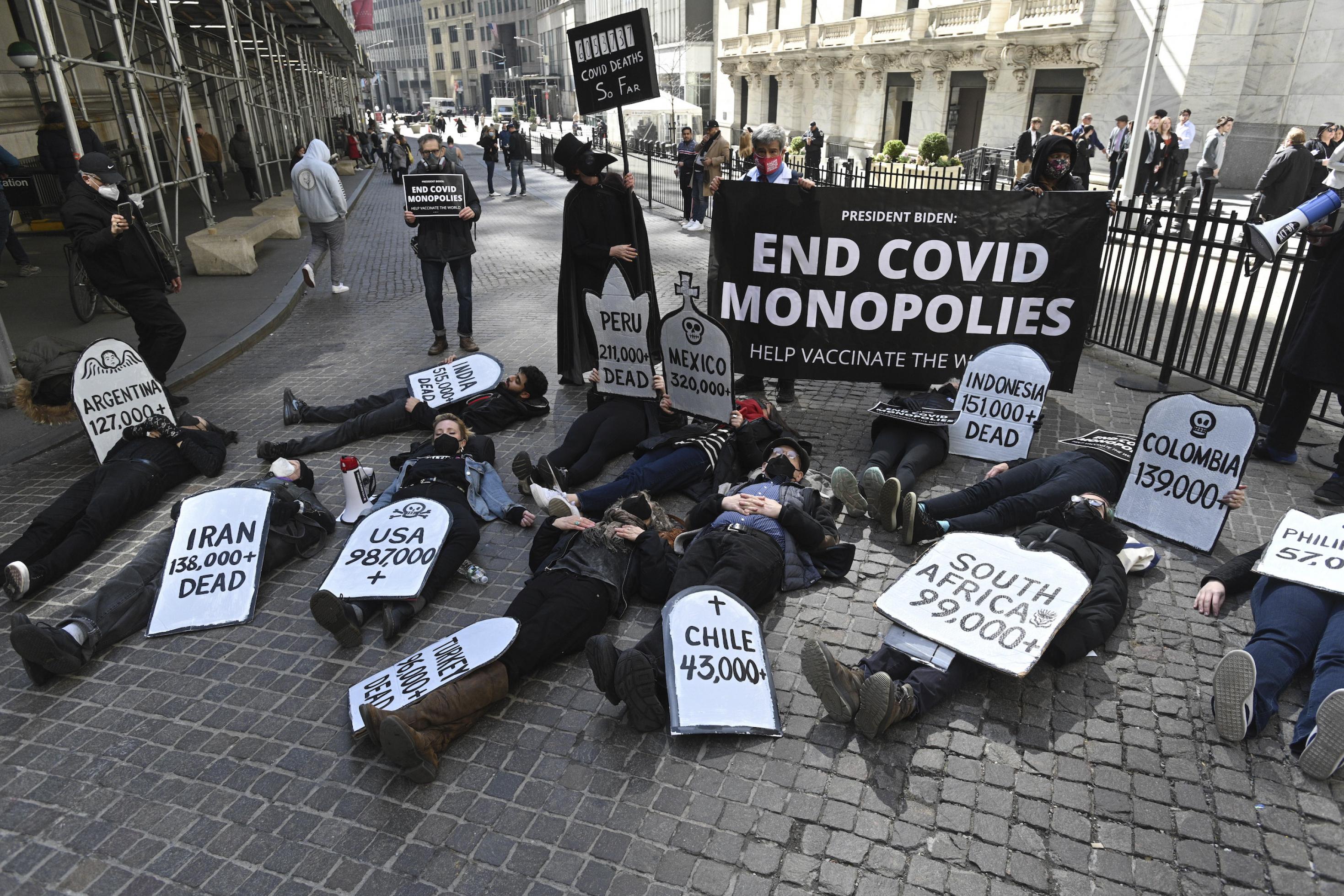 Etwa 20 Protestierende liegen auf dem Boden und halten Schilder fest, auf denen Ländernamen mit Corona-Todeszahlen stehen. Dahinter ein Plakat: „End Covid Monopolies“
