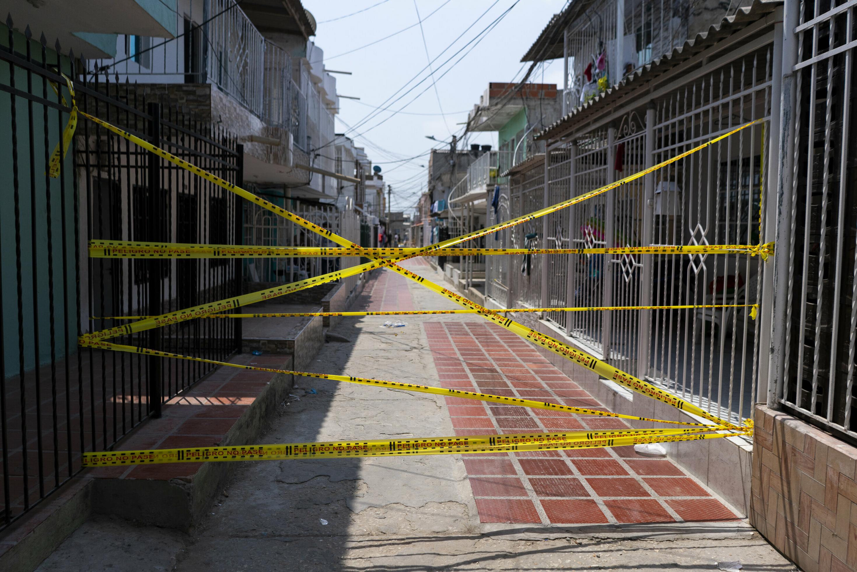 Das Foto zeigt den mit gelbem Warn-Plastikband kreuz und quer gesperrten Eingang zu einer Gasse in der kolumbianischen Karibikstadt Barranquilla. Die zweistöckigen Häuser stehen sich eng gegenüber. Sie grenzen sich zur Gasse mit mannshohen Gitterzäunen ab, was an einen Gefängnisflur erinnert.