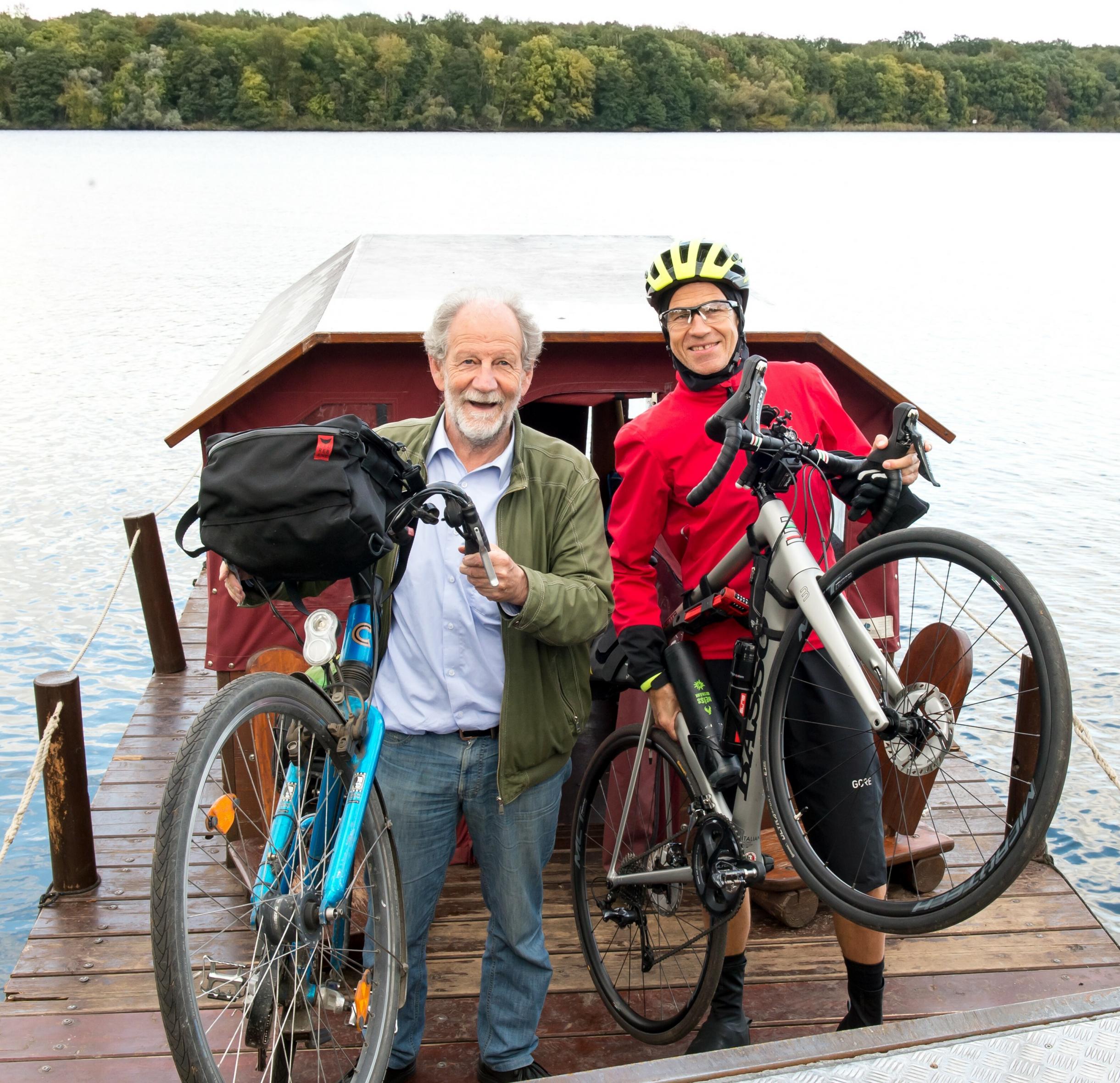 Michael Cramer und Martin C Roos lupfen nebeneinander lachend ihr Fahrrad am Bug des Wasservehikels; im Hintergrund der von der Havel gespeiste Jungfernsee.