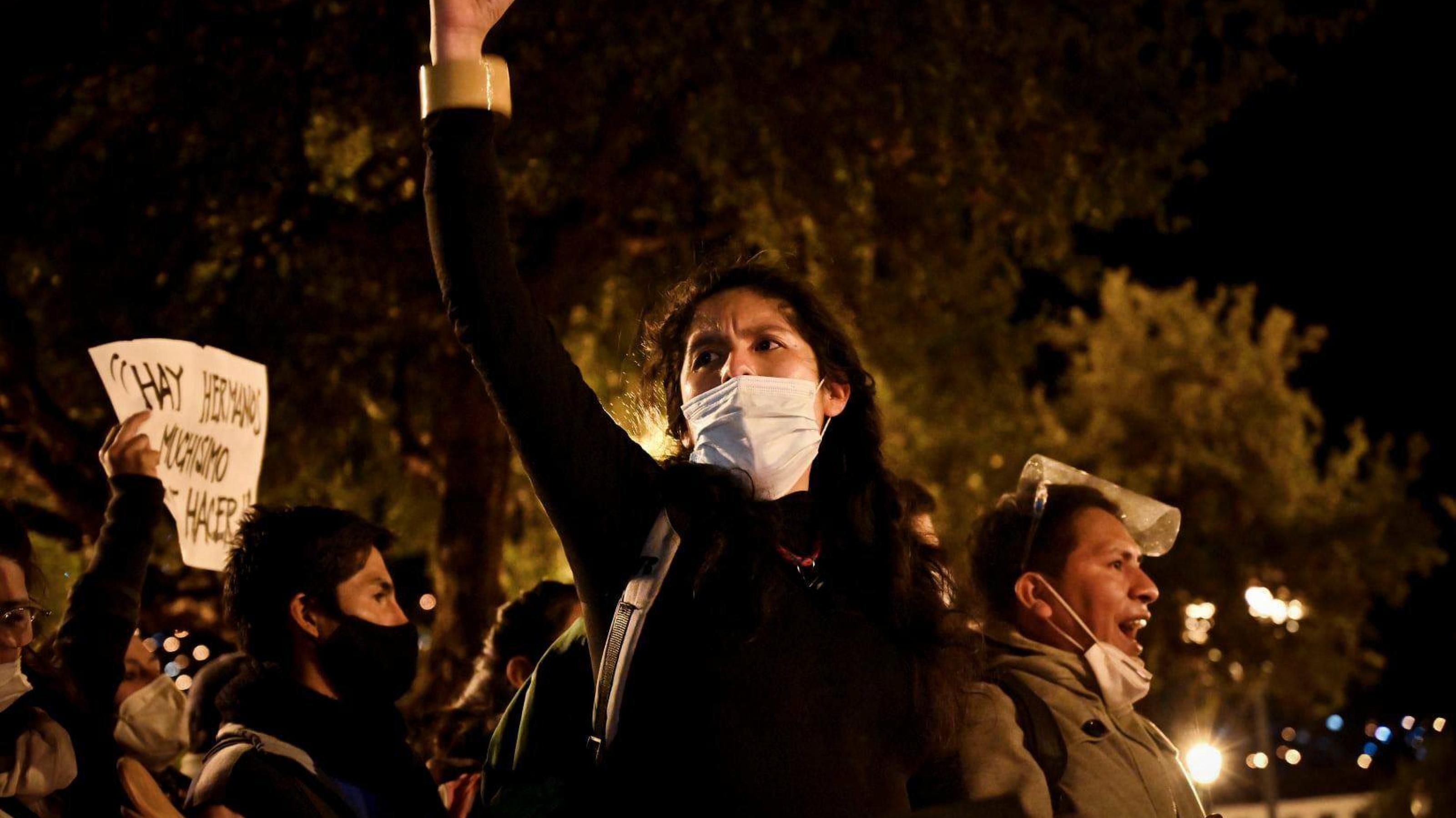 Junge schwarzgekleidete Frau mit weißem Mundschutz hält Hand kämpferisch in die Höhe. Es ist Nacht. Im Hintergrund Bäume, Laternen und weitere Demonstrierende.