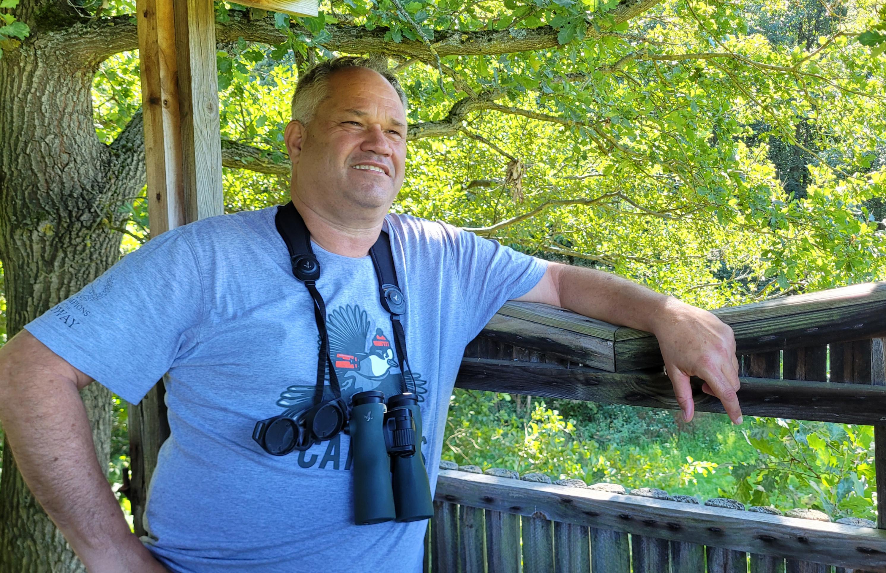 Dan Alon mit Fernglas um den Hals auf einem Hochsitz stehend, in kurzärmigen T-Sirt