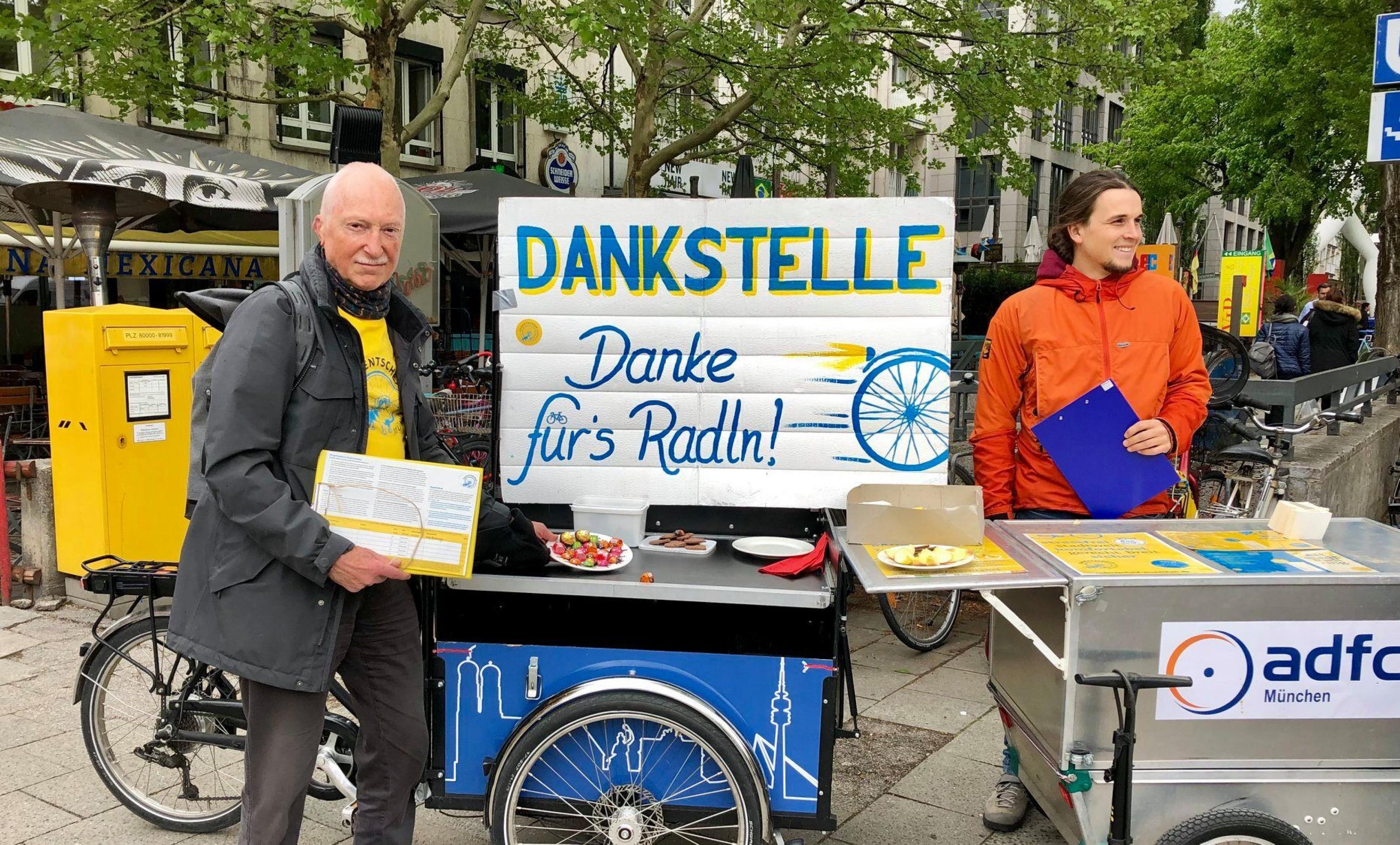Zwei Aktivisten des Münchner Radentscheid stehen am Rand eines Radwegs und verteilen Kuchen und Schokolade, um sich bei vorbeifahrenden RadlerInnen für das Radfahren zu bedanken.