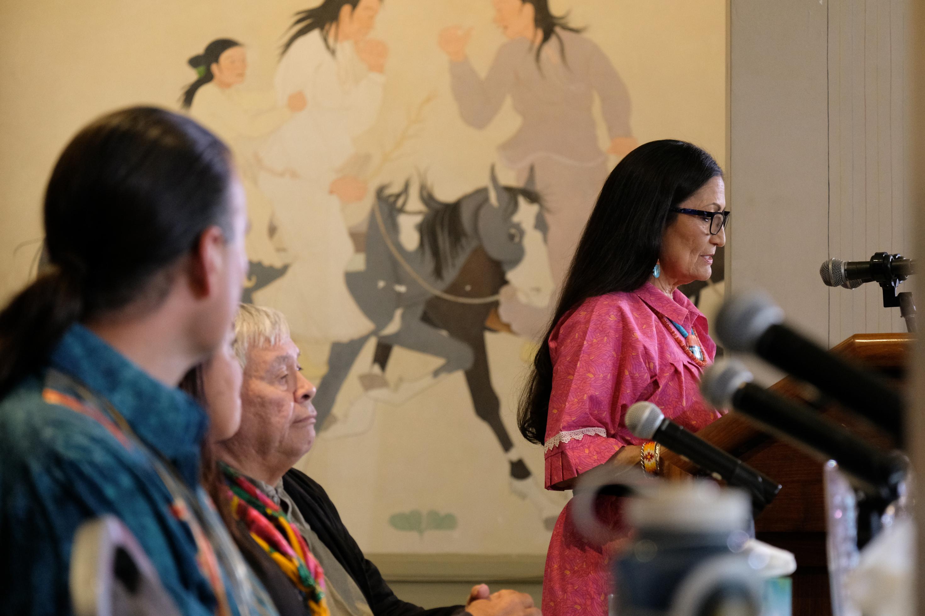 US-Innenministerin Deb Haaland steht in traditionellem pinkfarbenem Kleid am Podium., an dem Mikrofone angebracht sind. Im Hintergrund an der Wand ein indigener Wandschmuck. Stammesältere hören im Vordergund zu.