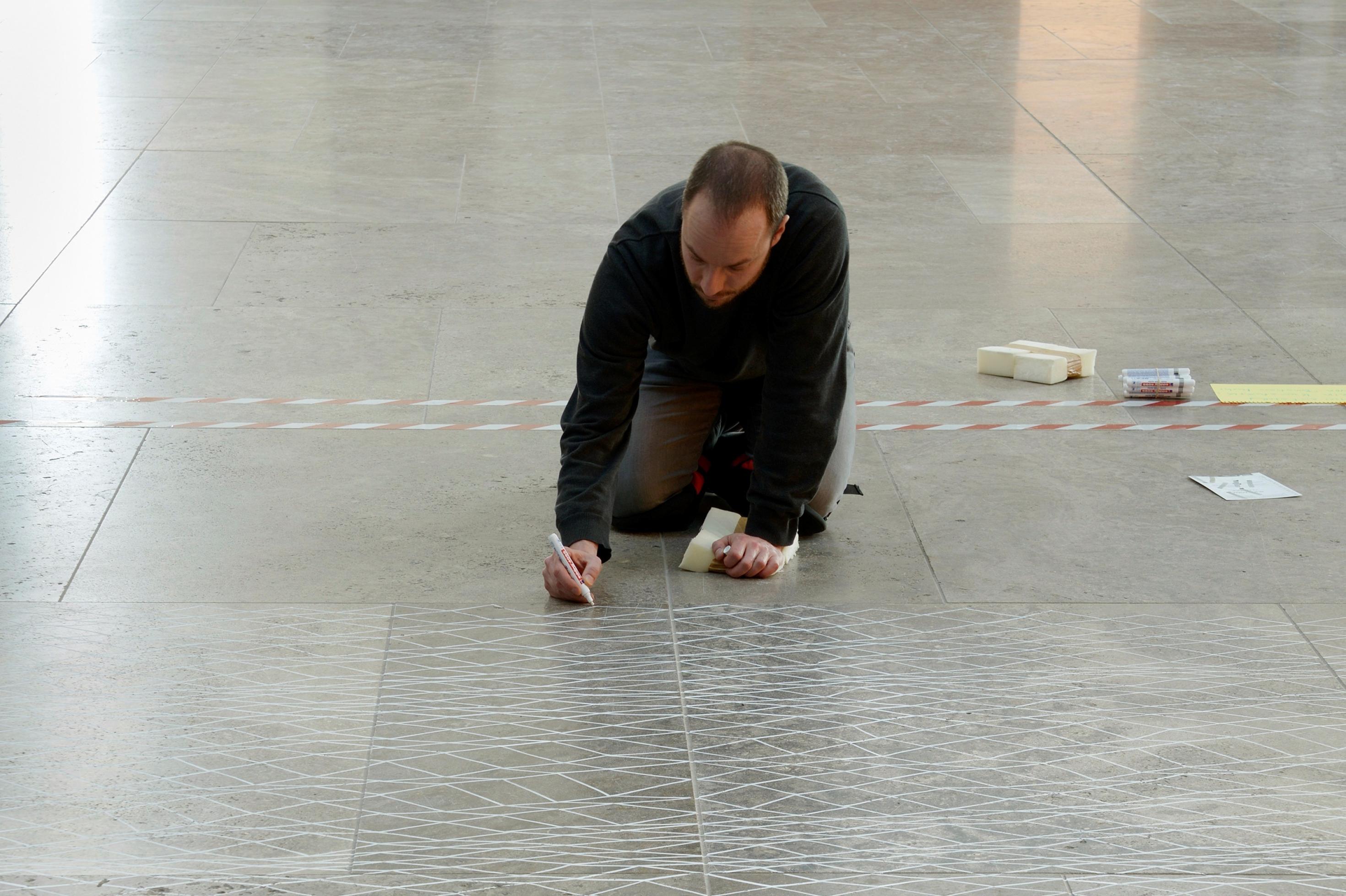 Ein Mann kniet auf dem grauen Boden und malt mit einem weißen Stift ein Netzmuster darauf.