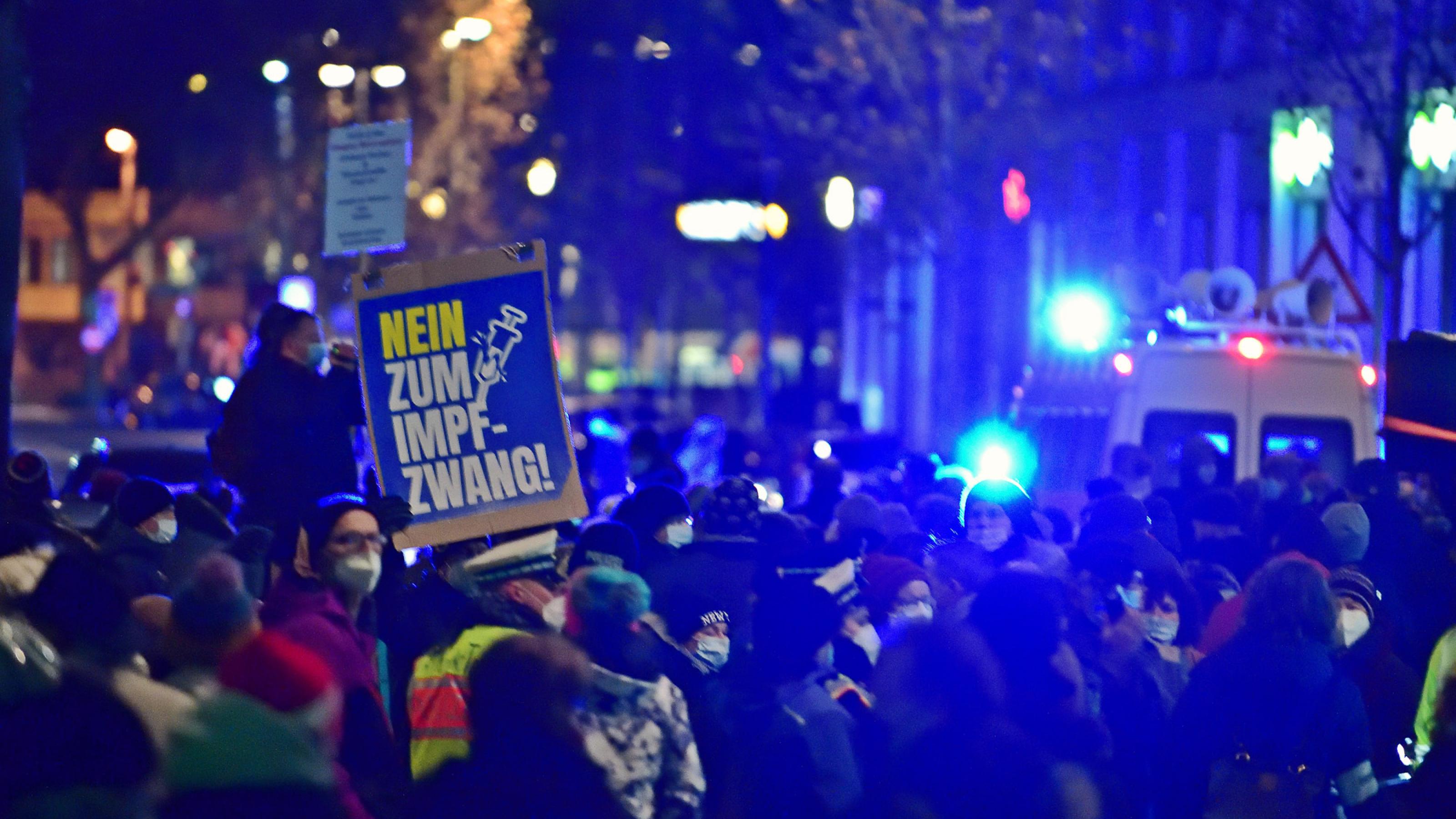 Eine Menschenmenge bei Nacht. Im Hintergrund ein Polizeiauto mit Blaulicht. Jemand hält ein Schild hoch mit der Aufschrift „Nein zum Impfzwang!“