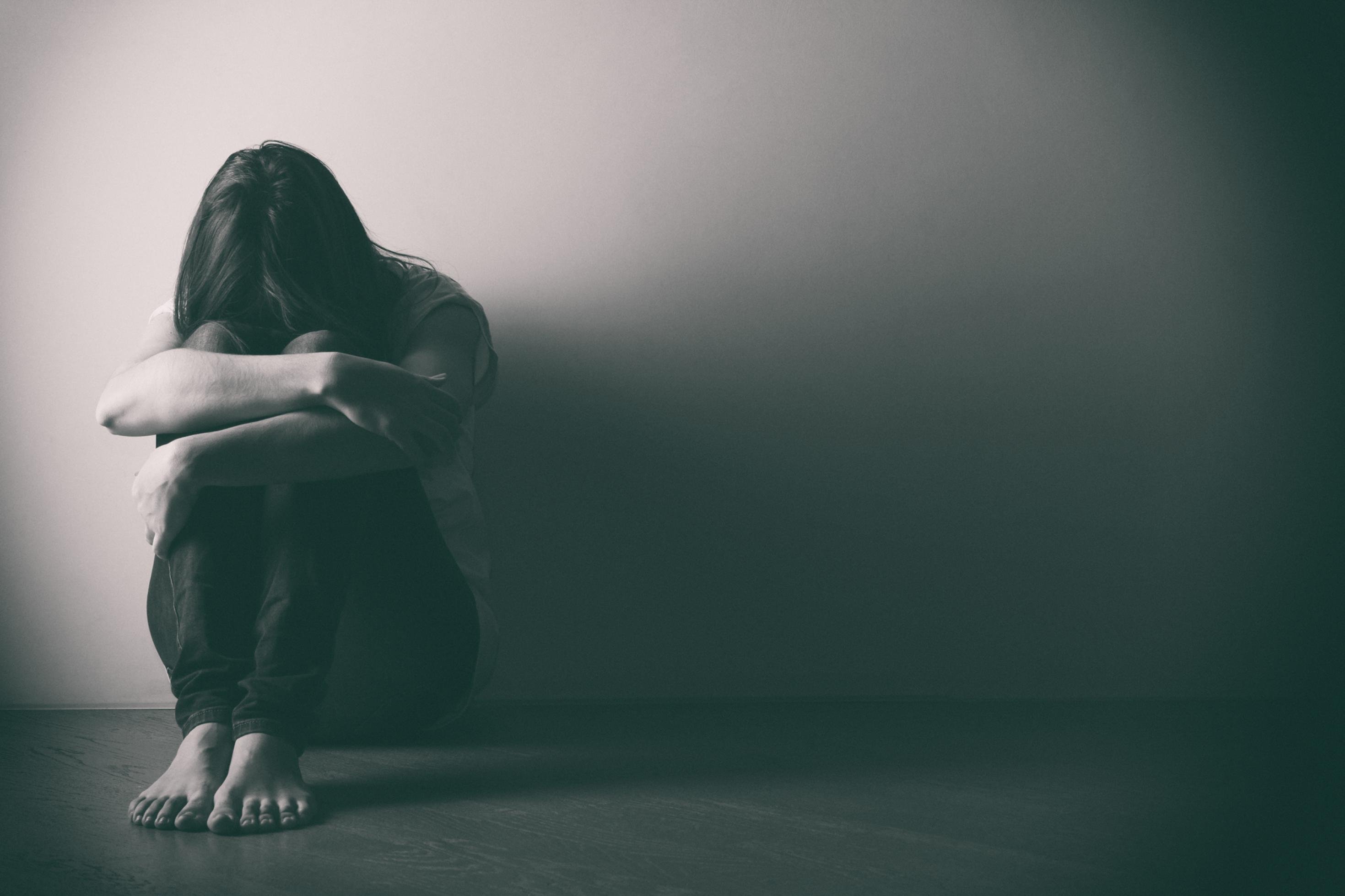 Auf dem Schwarz-Weiß-Foto ist eine Frau in lockerer Hose und T-Shirt zu sehen, die auf dem Fußboden vor einer kahlen Wand sitzt, die Knie angezogen, die Arme darauf verschränkt und das Gesicht nach unten geneigt, so dass nur ihre Haare zu sehen sind.