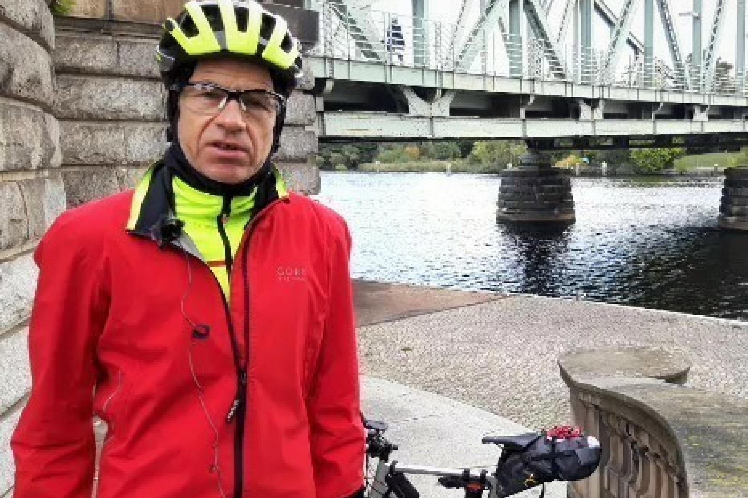 Im Vordergrund der Reporter in Radmontur und mit Radhelm, im Hintergrund sein Rennrad vor der Brücke mit der Metallkonstruktion über dem dahin fließenden Wasser der Spree.