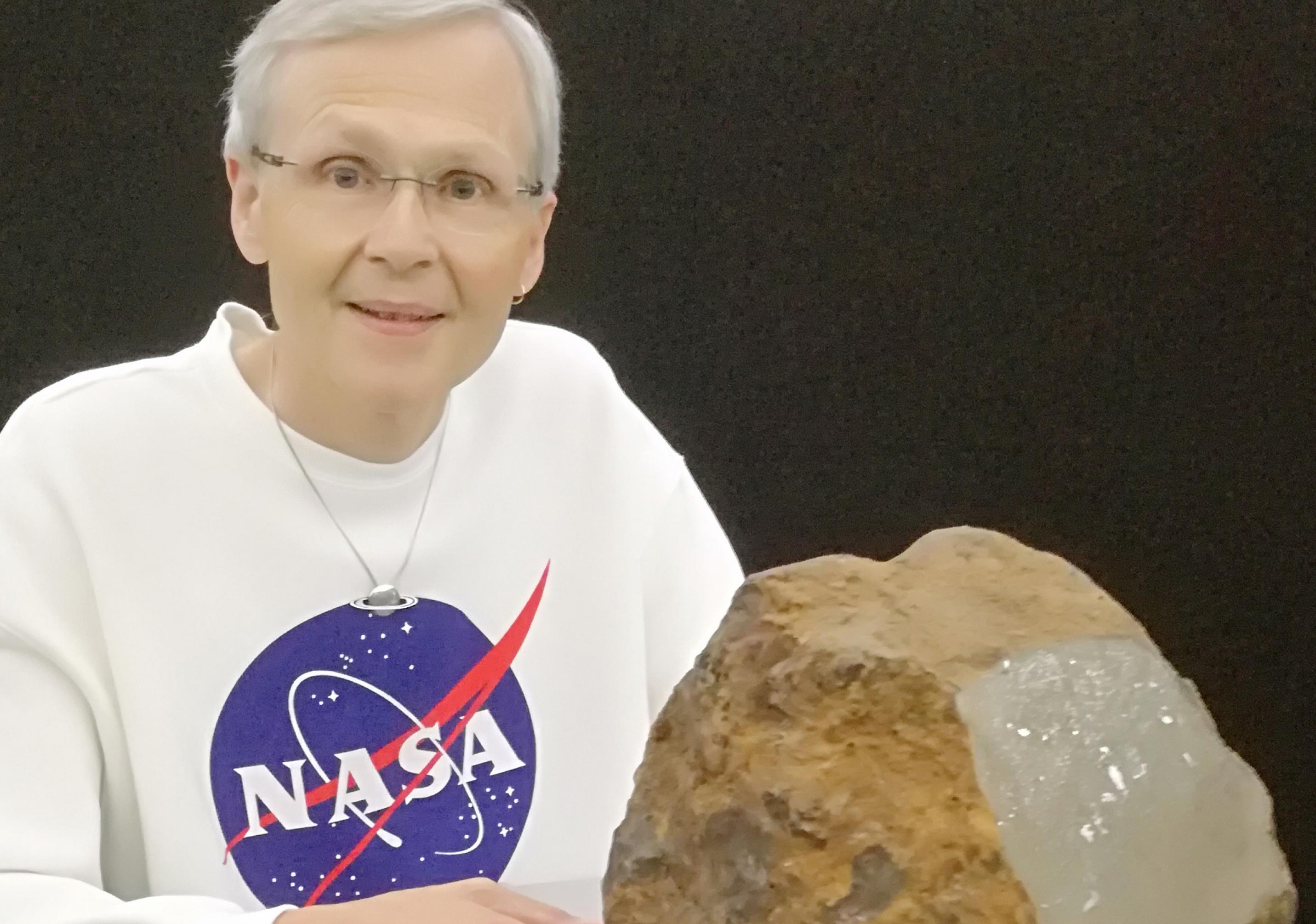 Meteoritenforscher Dieter Heinlein mit einem weißen Pullover mit NASA-Logo. Vor ihm liegt der rucksack-große und rost-organene Meteorit aus Blaubeuren.