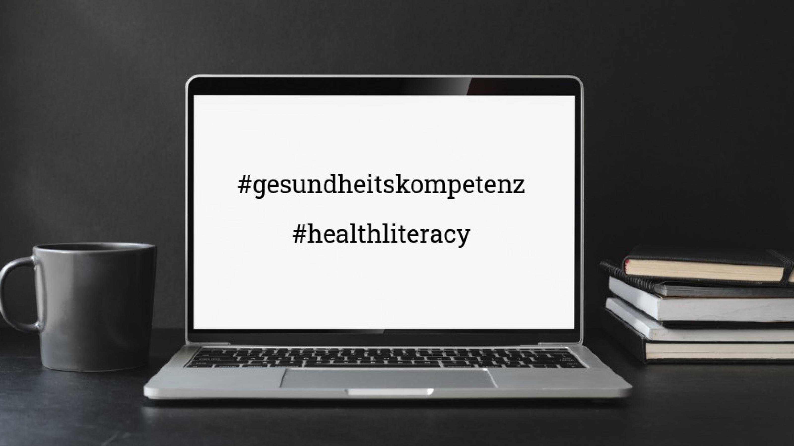 Zwischen einer Tasse auf der linken Seite und einem Bücherstapel auf der rechten Seite steht ein aufgeklappter Laptop. Auf dem Screen sind die Hashtags #gesundheitskompetenz und #healthliteracy zu sehen.