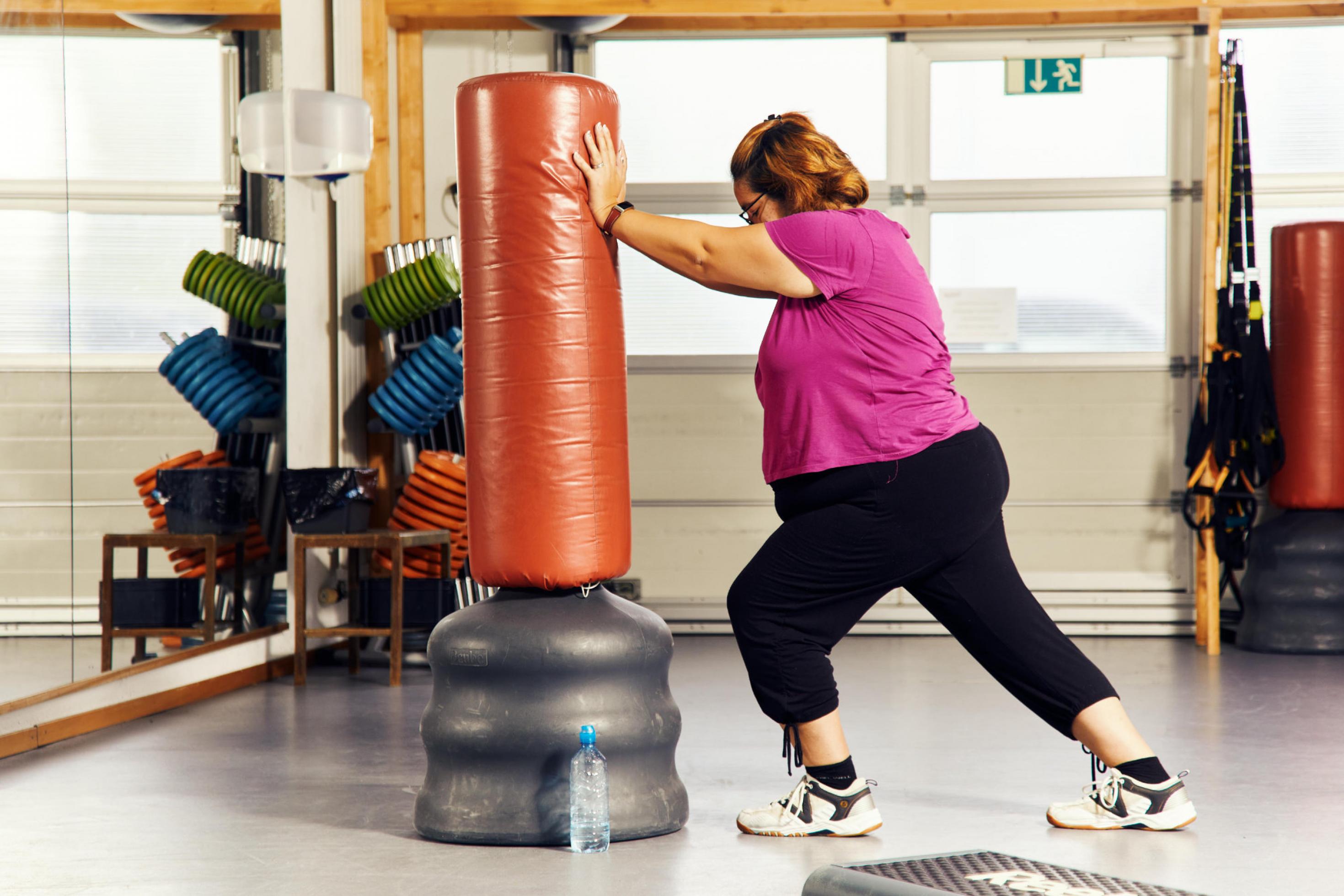 Eine stark übergewichtige Frau trainiert in einem Gymnastikraum mit einem stehenden Boxsack.