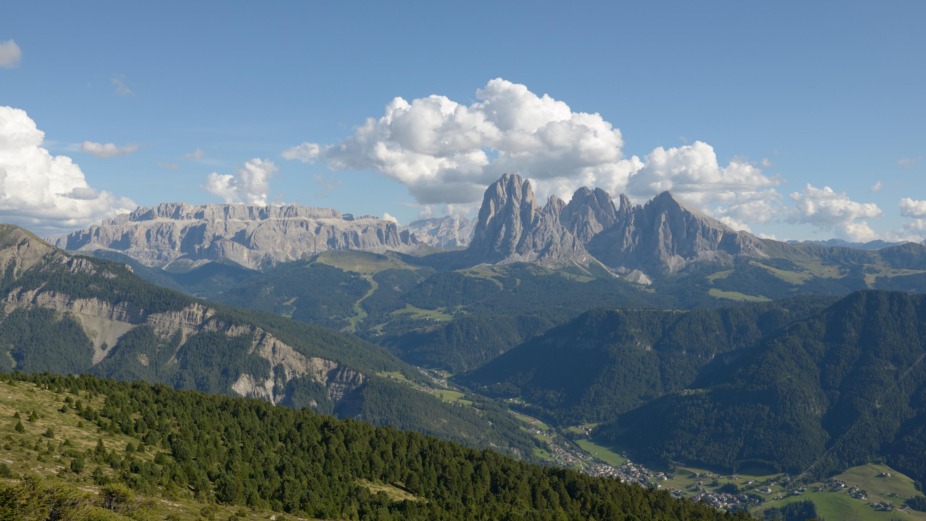 Ein Bergpanorama, in der Ferne schroff aufragende Dolomitenberge unter weißen Wölkchen, im Vordergrund Wälder und Wiesen
