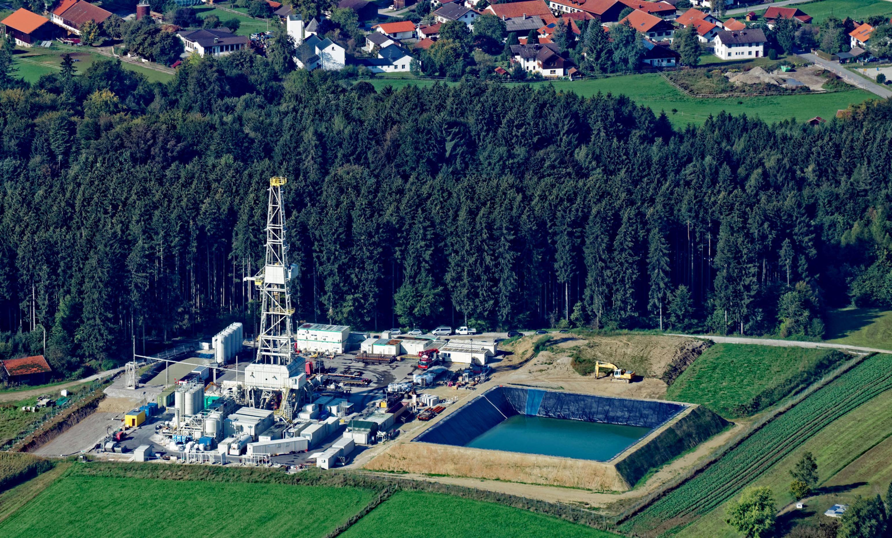 Luftbild: oben ein Dorfrand, darunter ein Waldstreifen, darunter ein Bohrturm mit einigen Baucontainern, Zementbehältern und einem Wasserbecken. Weiter unten schließen sich grüne Felder an.