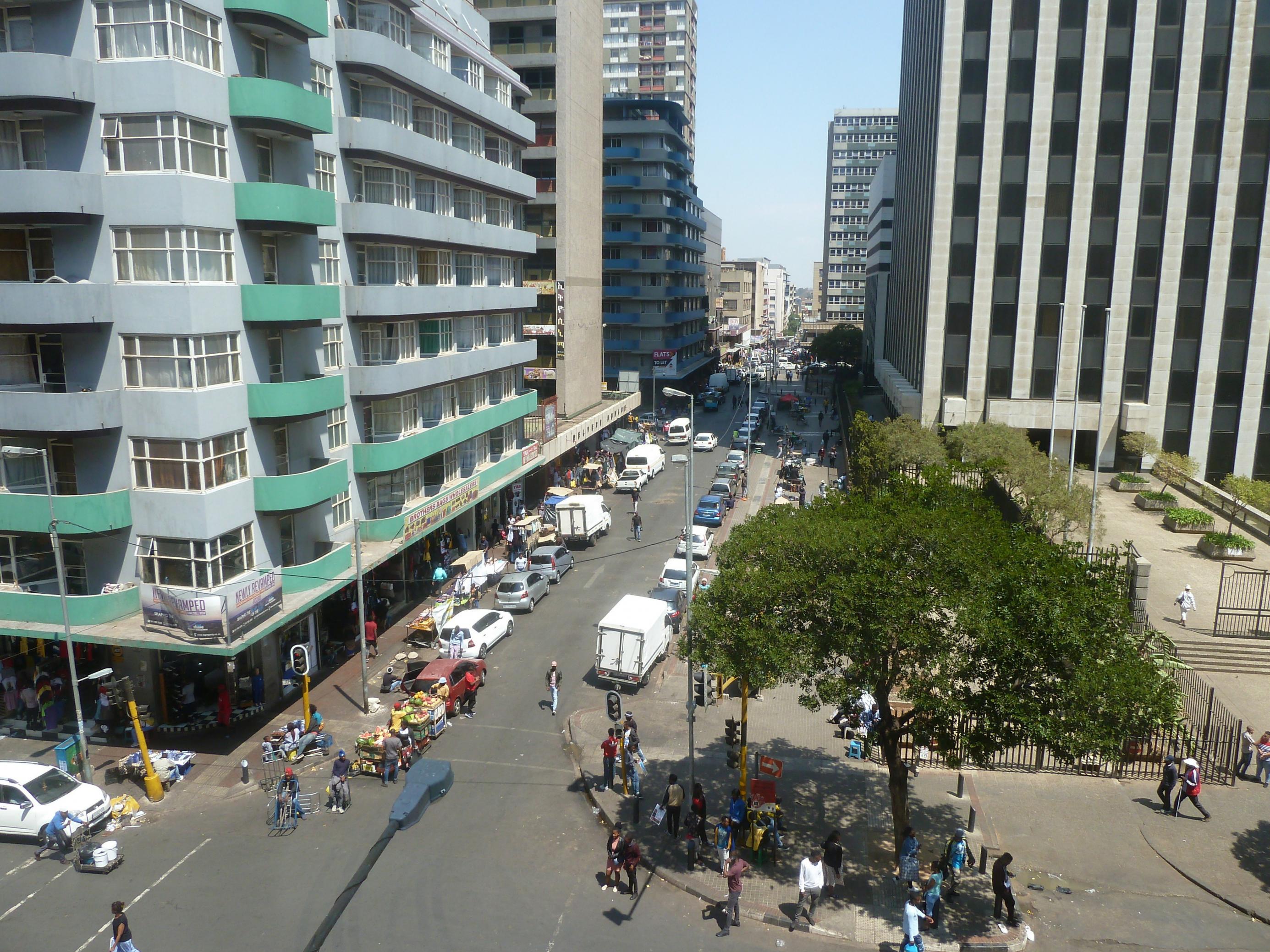 Eine Straßenschlucht im Zentrum von Johannesburg, an den Straßenrändern parken Autos, Fusßgänger sind unterwegs. Vorne rechts im Bild wachsen ein paar Bäume.