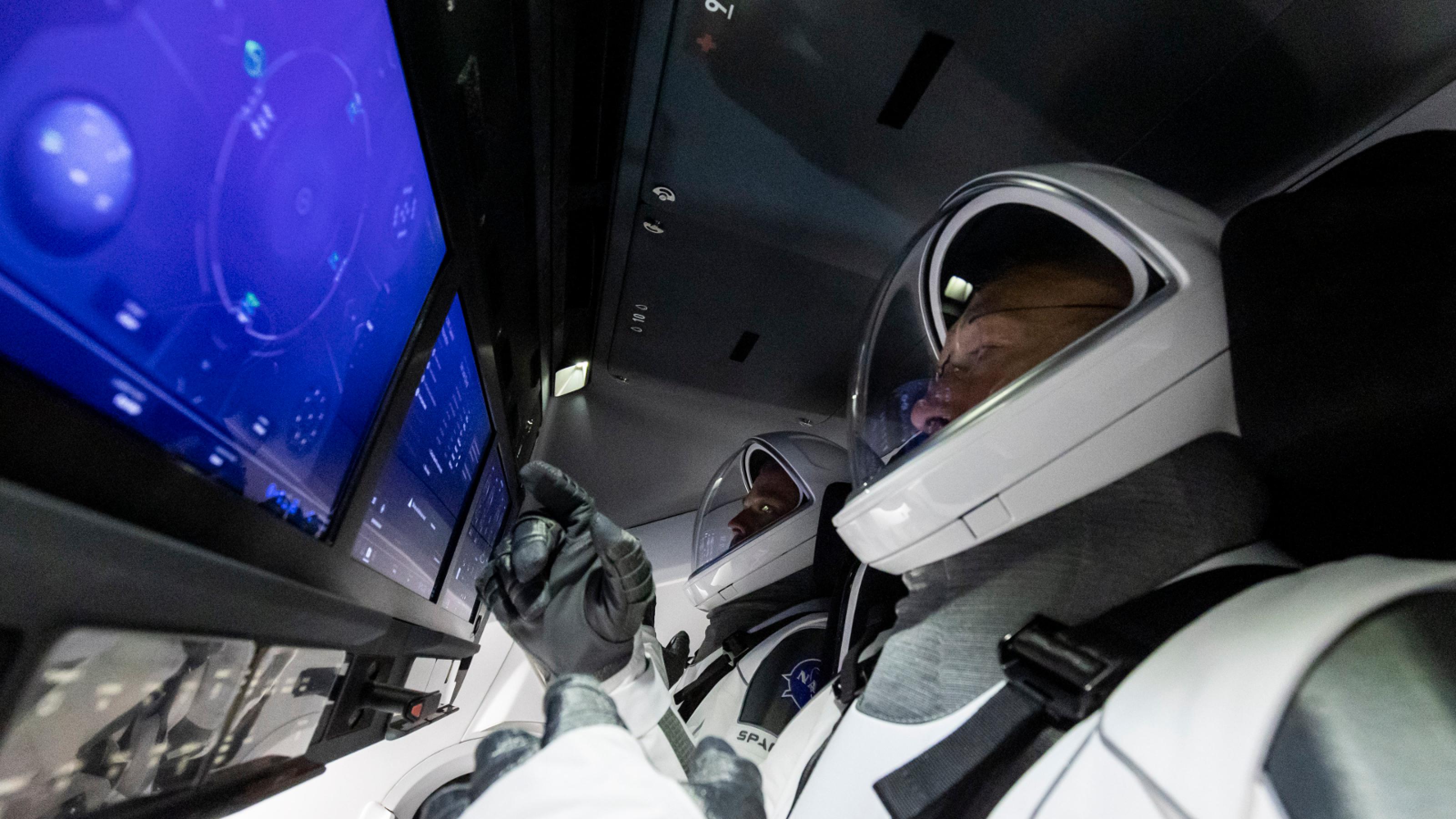 Zwei Astronauten, deren Helme man sieht, blicken auf blau schimmernde Touchscreens im Cockpit der Kapsel Dragon-2