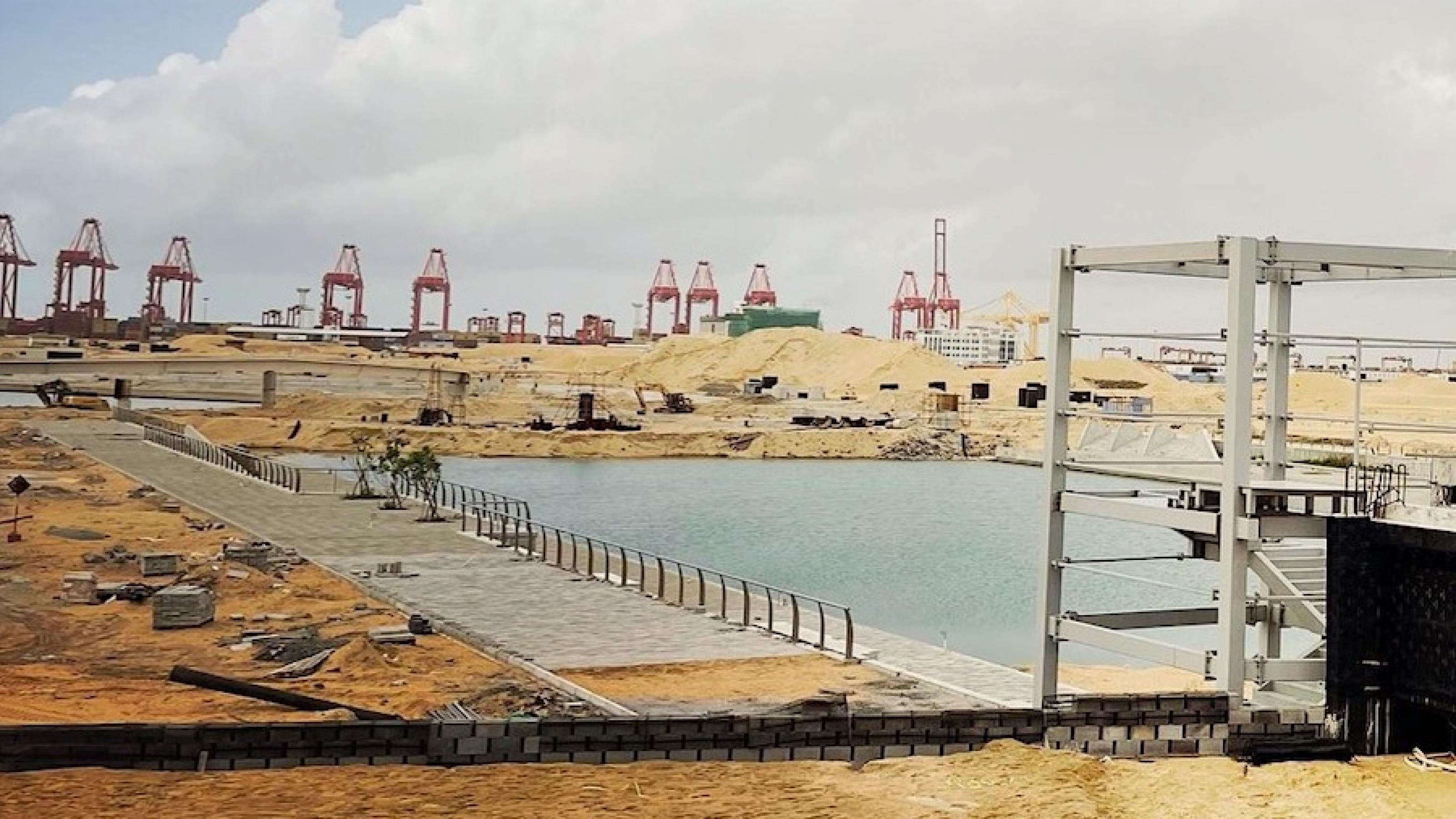 Sandige Großbaustelle in einem Hafen. Im Vordergrund ist ein Wasserbecken zu sehen, im Hintergrund Hebekräne.