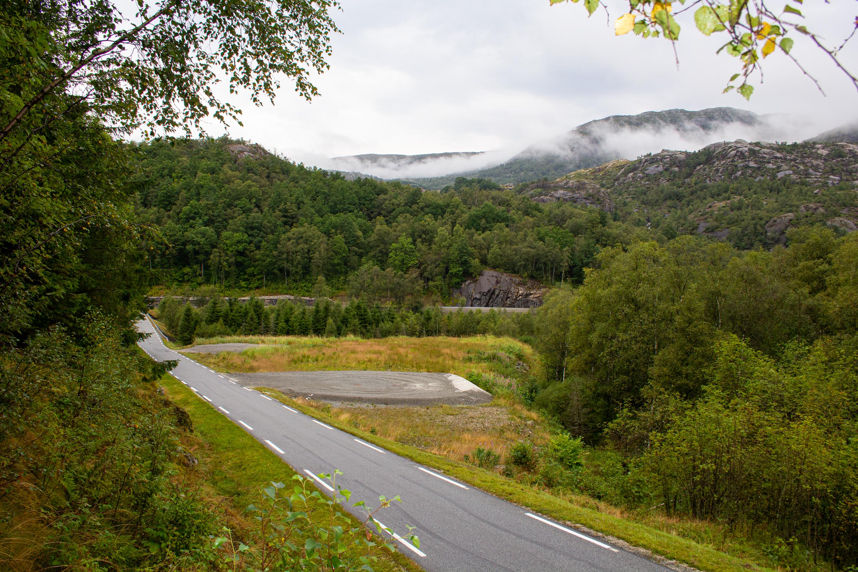 Schotterfläche am Straßenrand in bergiger Landschaft; aus dem Wald steigen Wolken auf