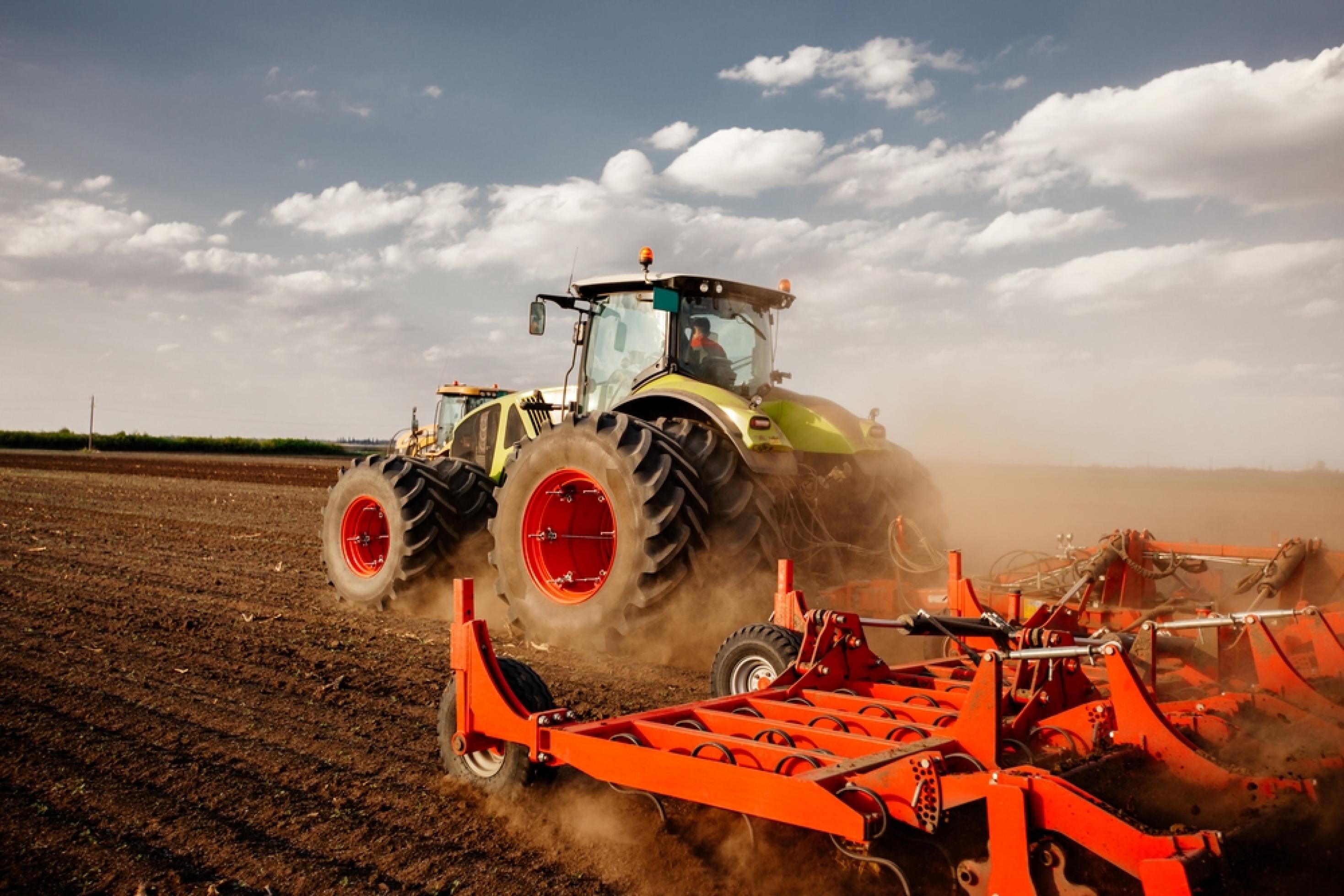 Ein Traktor zieht auf einem leeren Feld eine rote Landmaschine hinter sich her.