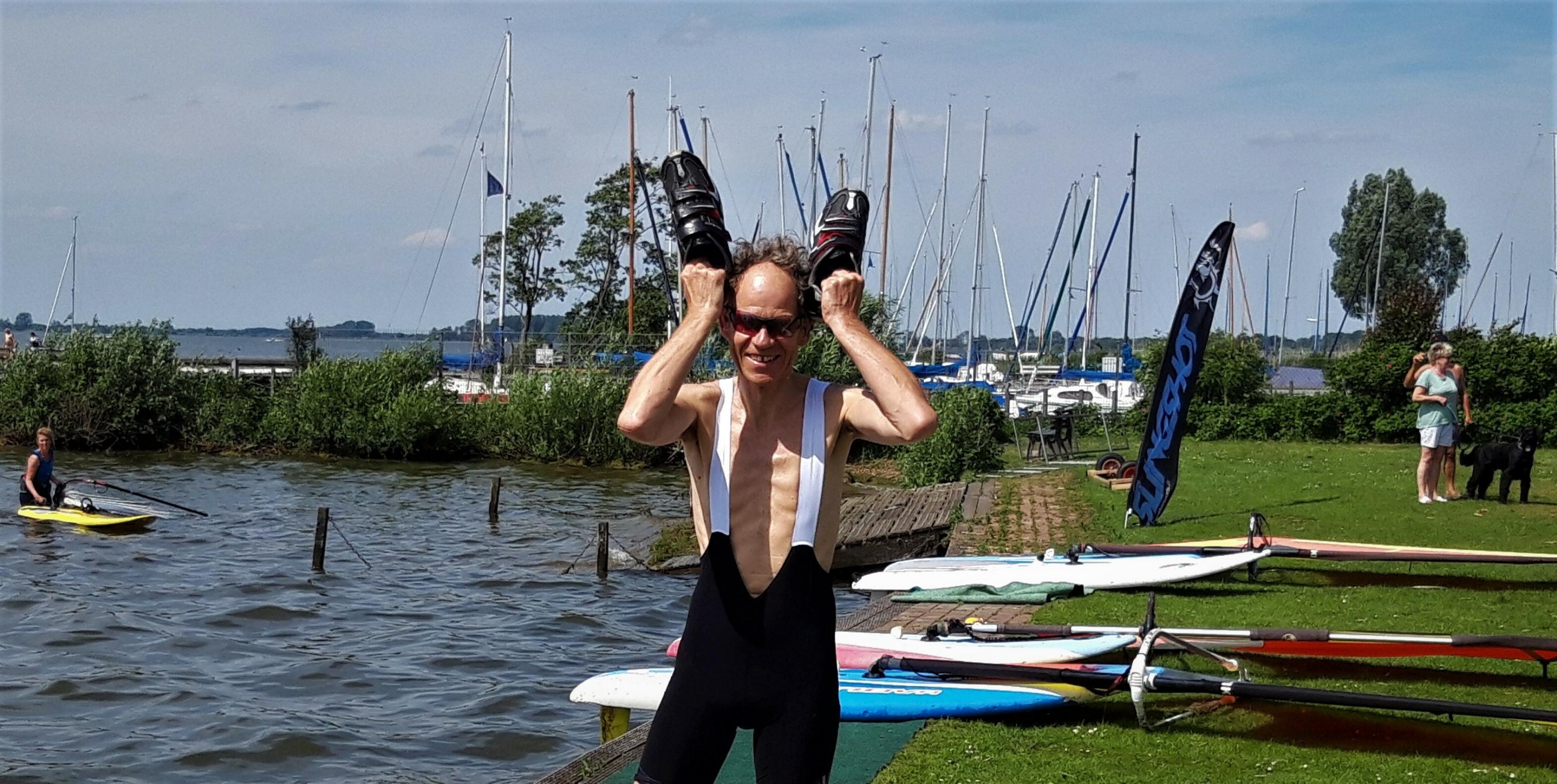 Autor Martin C posiert mit den Radschuhen in der Hand am vom Wind aufgewühlten Wasser des Sees.