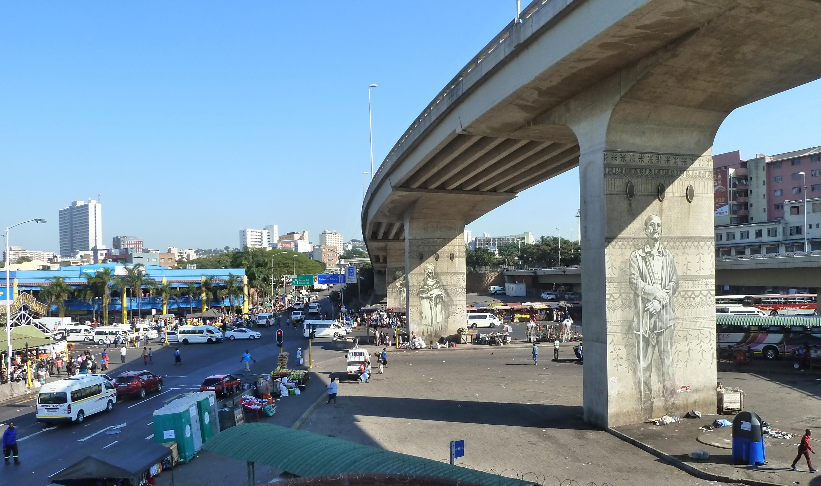 Eine Betonbrücke, auf deren Pfleiler große Porträts gemalt wurden, führt in die Innenstadt von Durban. Hochhäuser sind zu sehen, davor Straßenhändler und Minibus-Taxis.