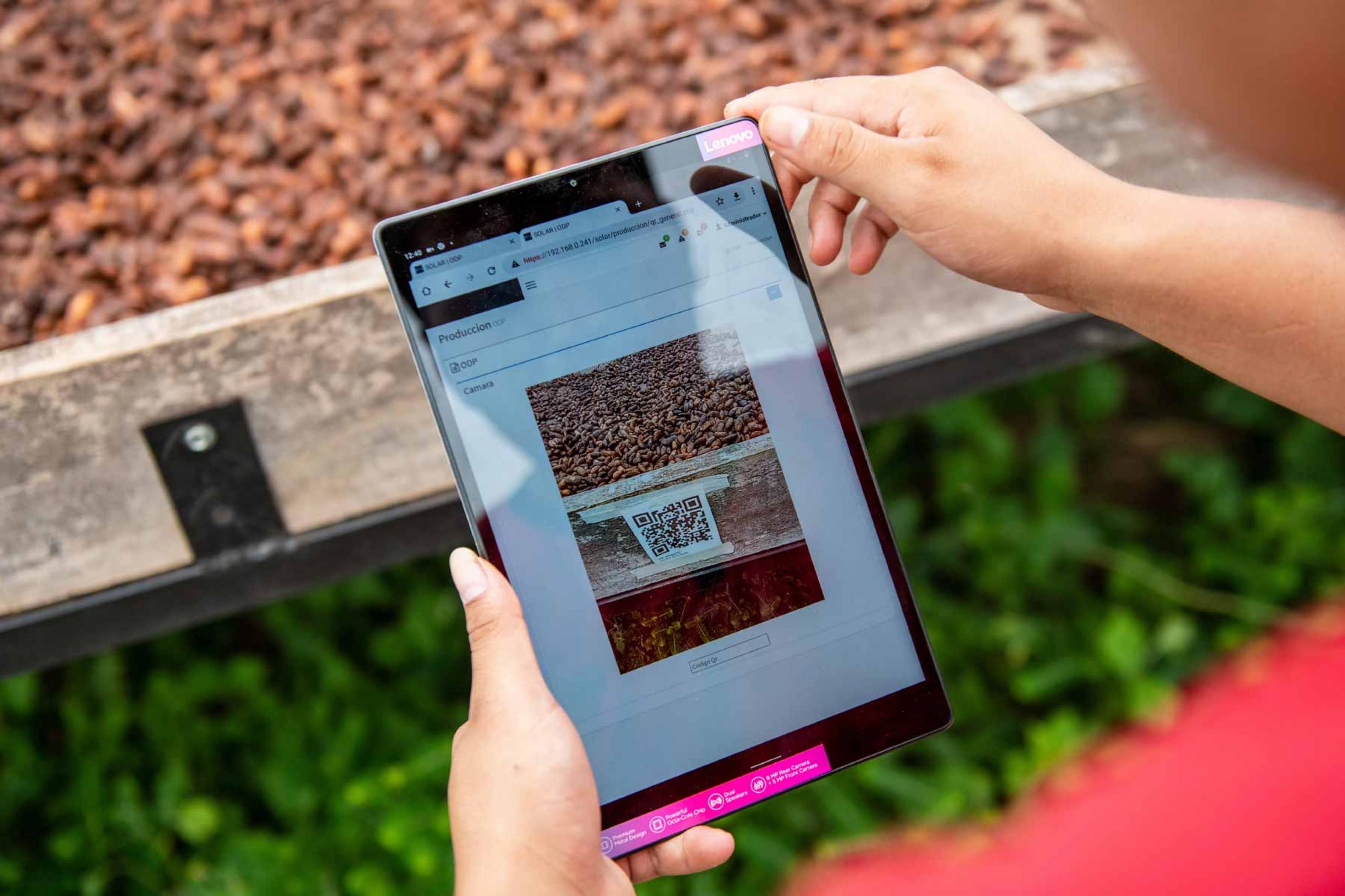 Ein Tablet im Vordergrund mit einem QR-Code, dahinter eine Holzkiste mit Kakaobohnen.