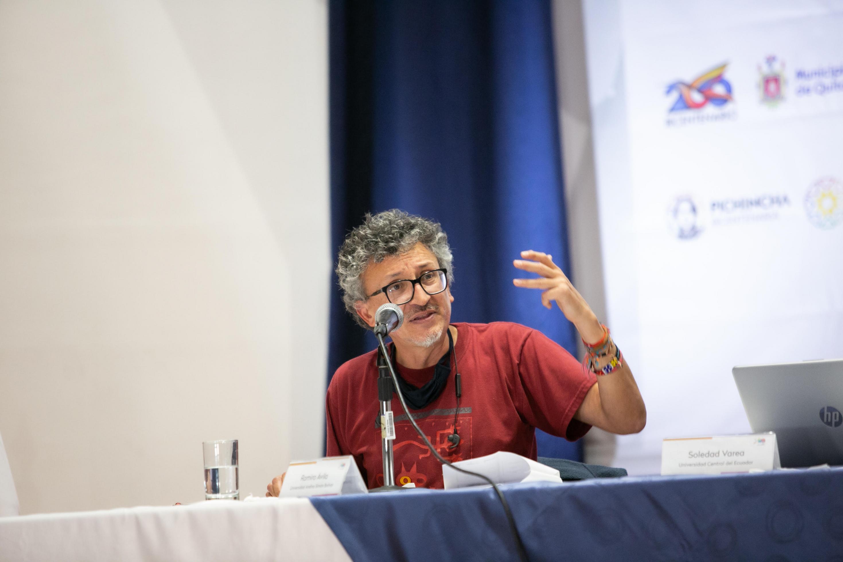 Ein Mann mit grauen Locken und einer Brille gestikuliert an einem Tisch während einer Podiumsdiskussion.