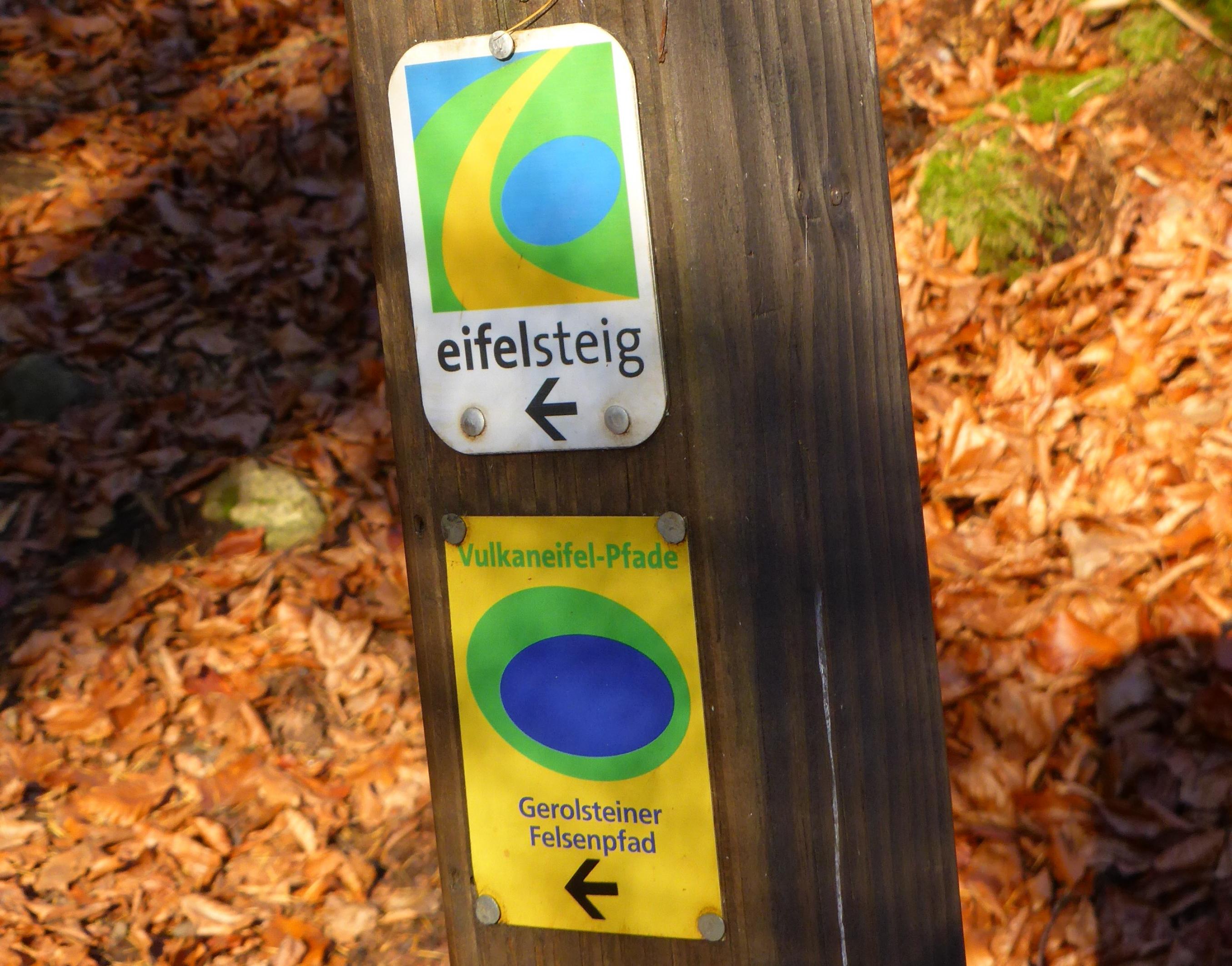 Wanderschilder an einem Holzpfosten, hinter dem rotgoldenes Herbstlaub in der Sonne leuchtet.