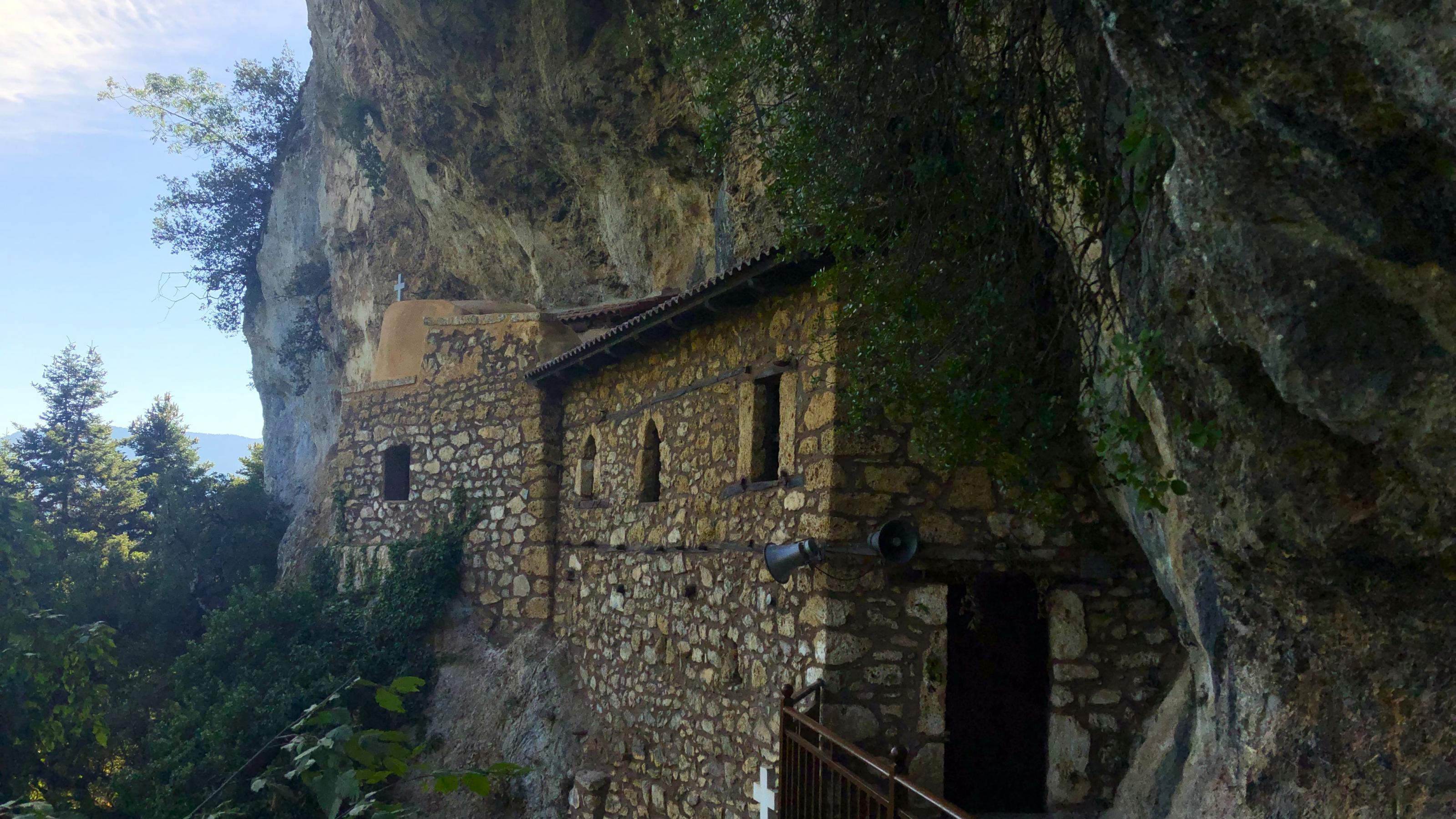 In einer steilen Felswand klebt unter einem Überhang ein einfaches Steinhaus an der Wand.