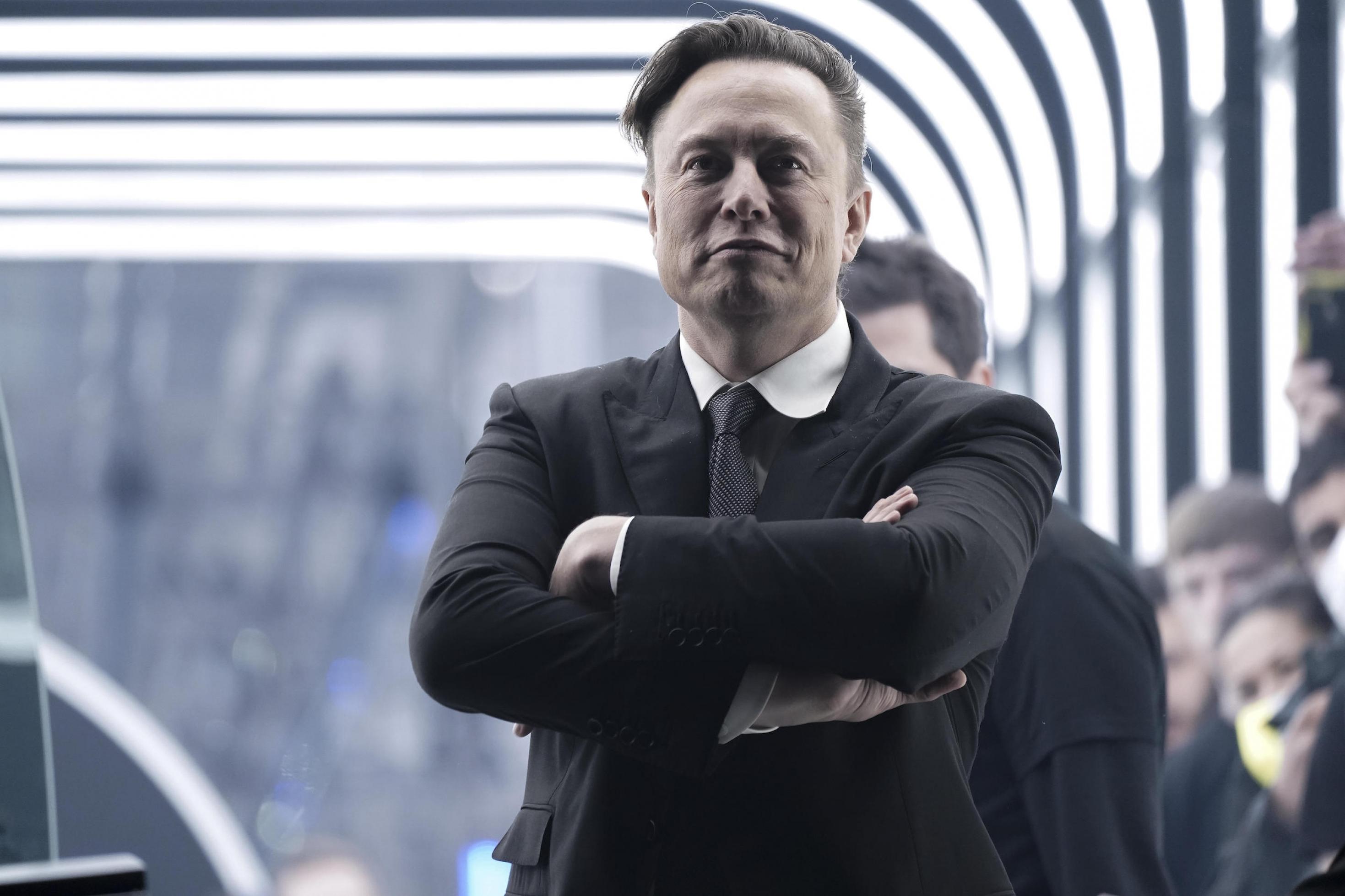 Musk, ein Mann mit markanten Gesichtszügen, steht mit verschränkten Armen und einem entschlossenen Gesichtsausdruck vor einer schwarz-weißen Kulisse