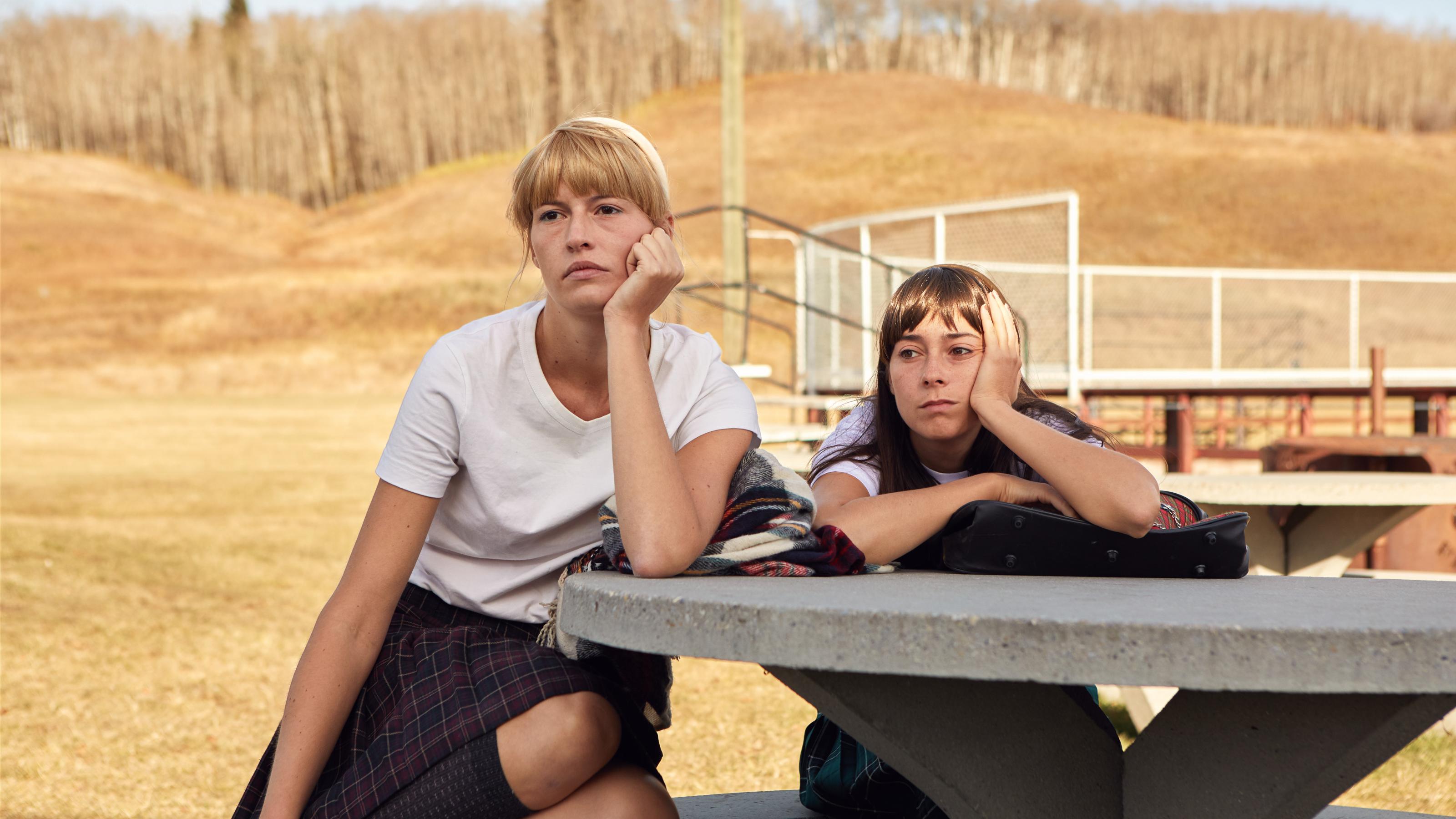 Zwei junge Frauen sitzen auf einem der Geräte eines leeren Spielplatzes und schauen in die Kamera.