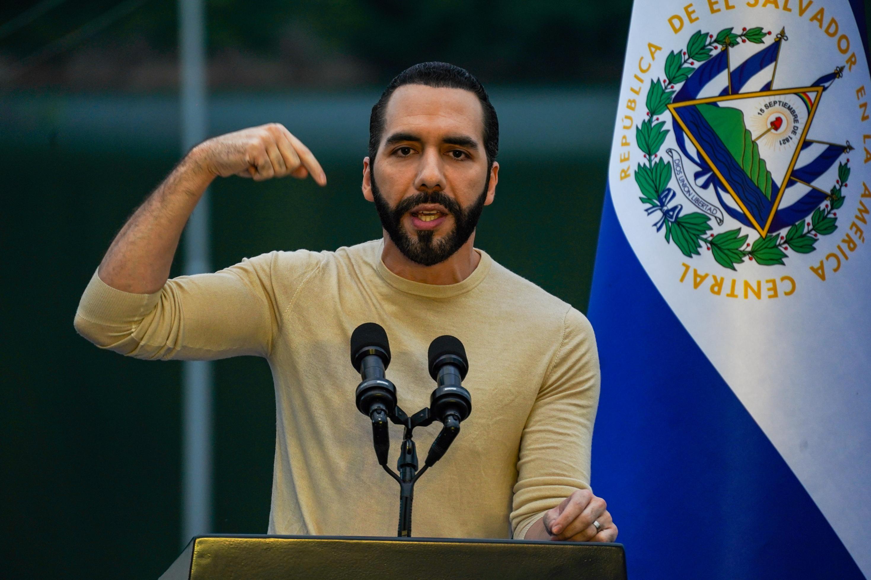El Salvadors Präsident Bukele im beigen, hautengen Pulli, gestikuliert an einem Rednerpult, dahinter die Landesflagge.