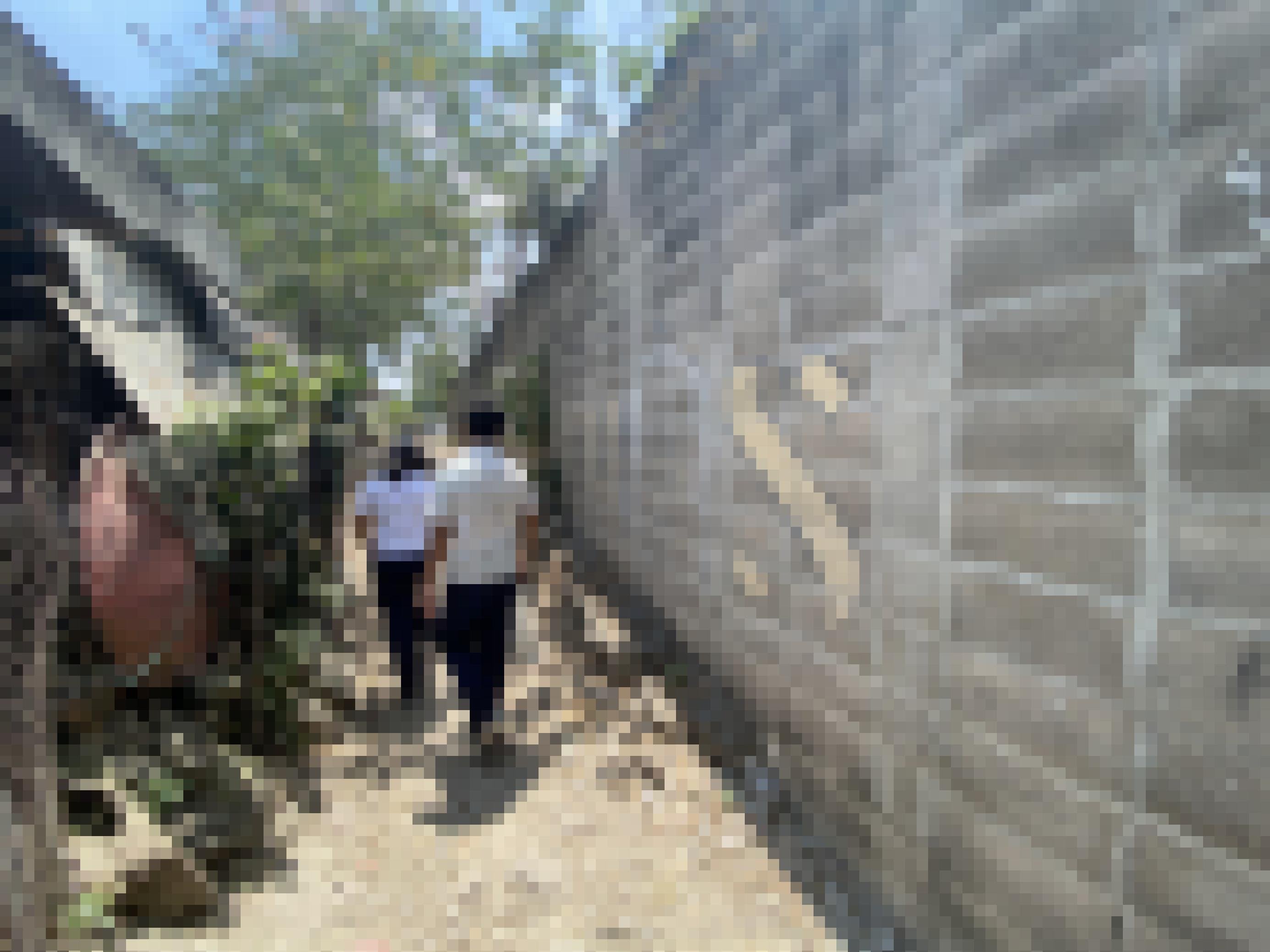 Eine unverputzte Mauer mit einem Graffiti MS, daran laufen zwei Schüler in weisser Uniform vorbei auf einem Trampelpfad.