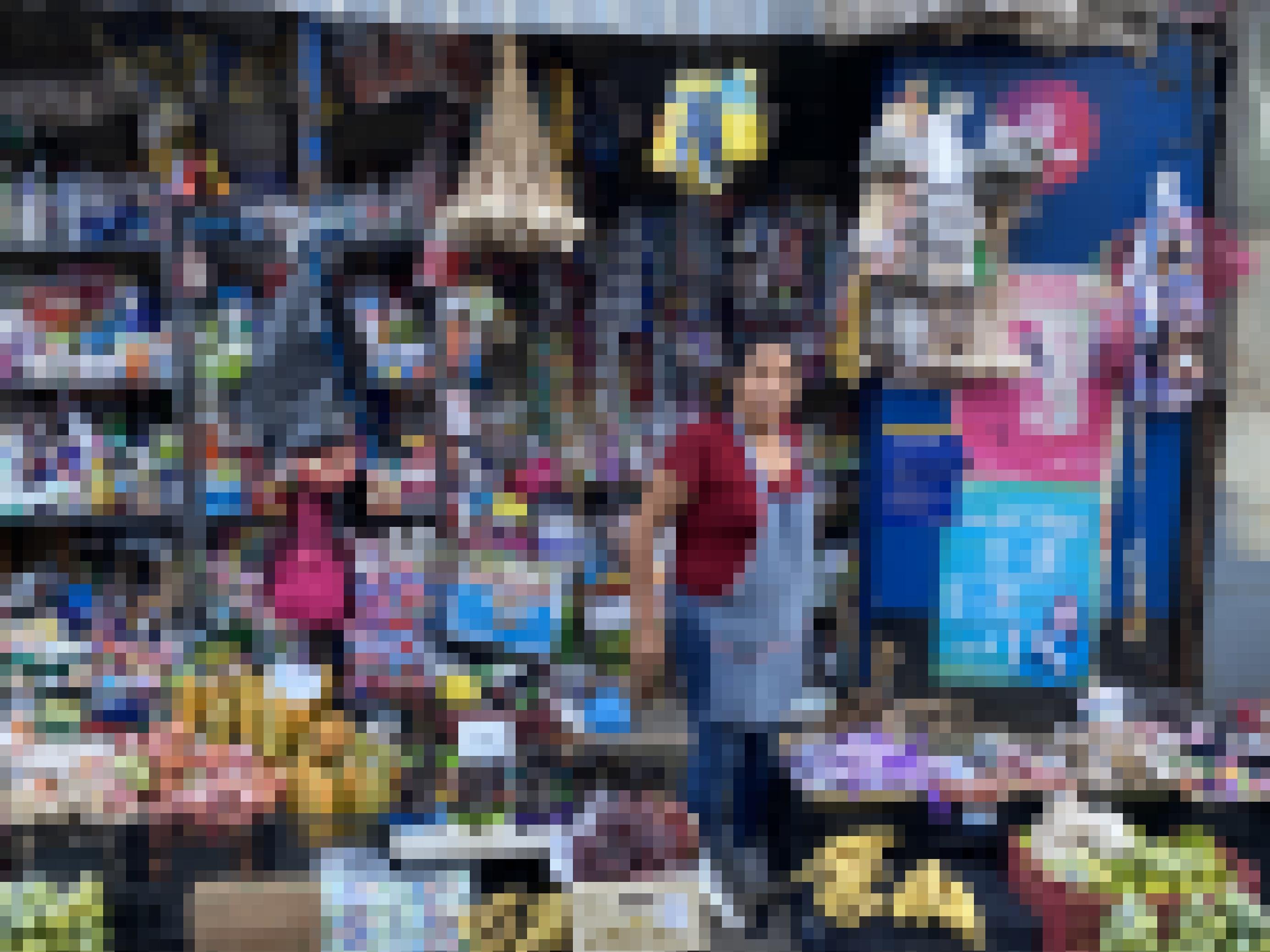 Eine Frau mit einer Schürze umgeben von einem bunten Durcheinander mit Waren aller Art, von Obst bis Medikamenten.