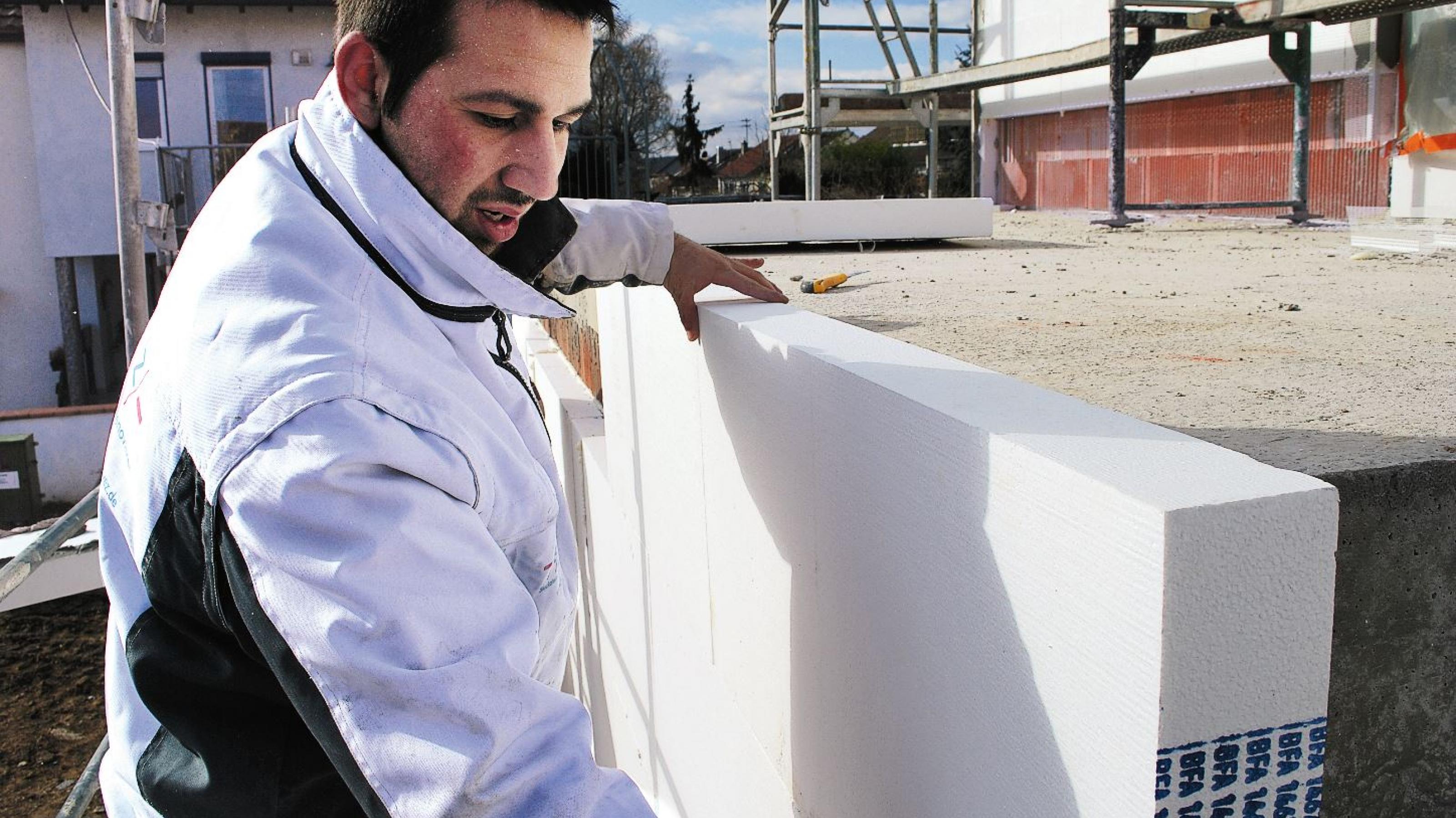Ein Handwerker bringt eine Dämmplatte an einer Betonwand an, im Hintergrund ein Baugerüst.