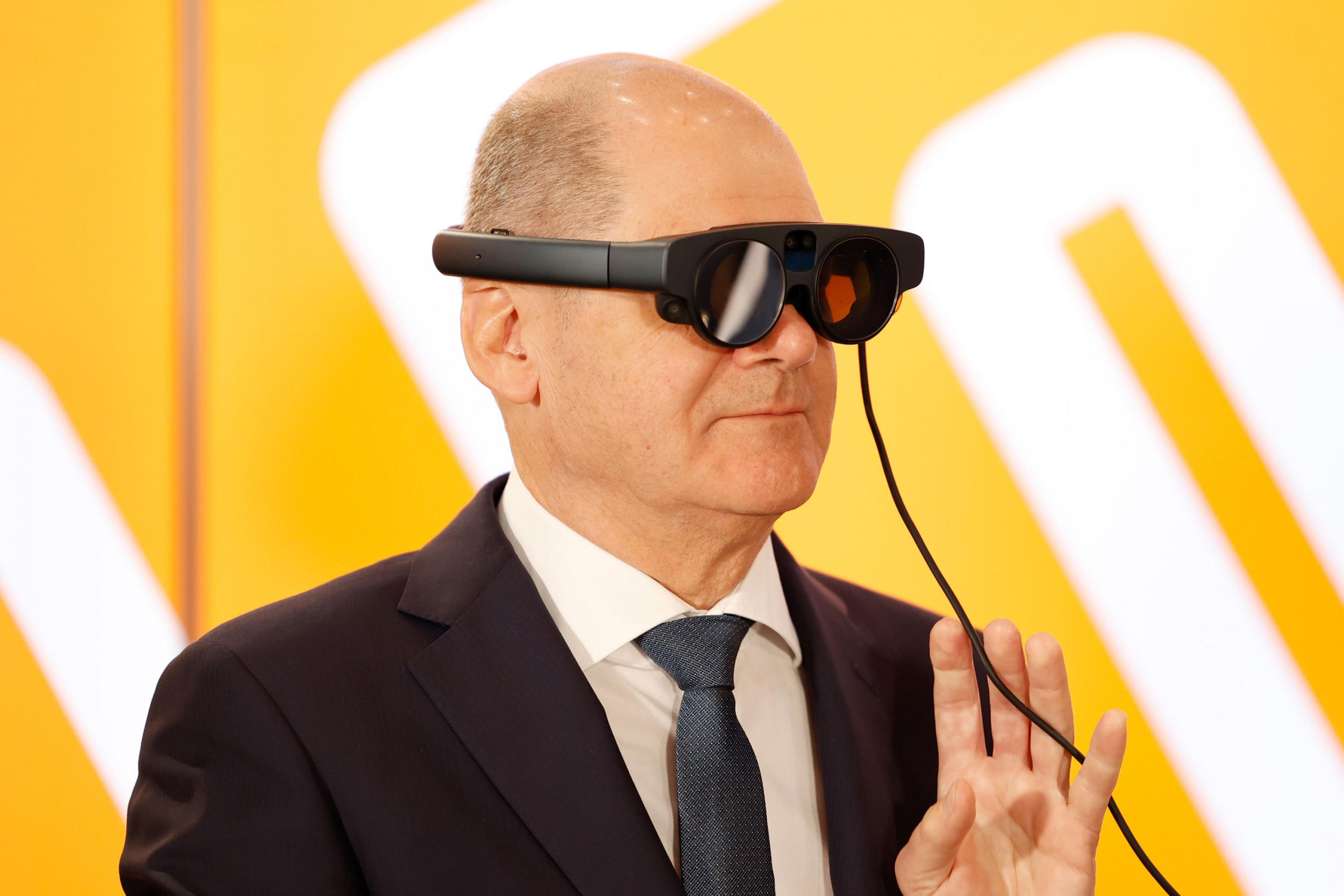 Kanzler Scholz mit VR-Brille vor gelbem Hintergrund.