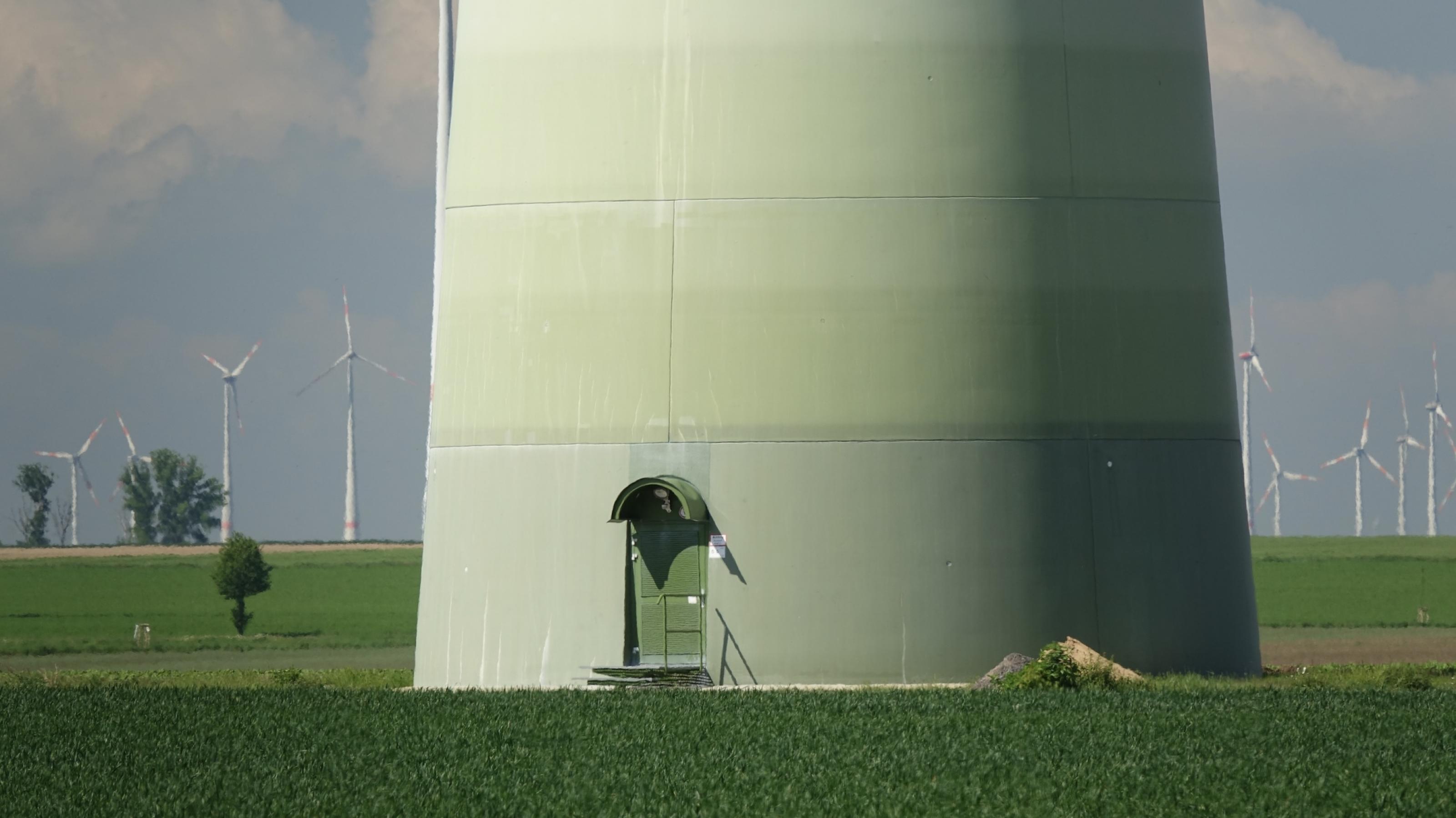 Ein riesiger Turm eines Windkraftwerks mit grün gestrichener Eingangstür, am Horizont weitere Windräder.