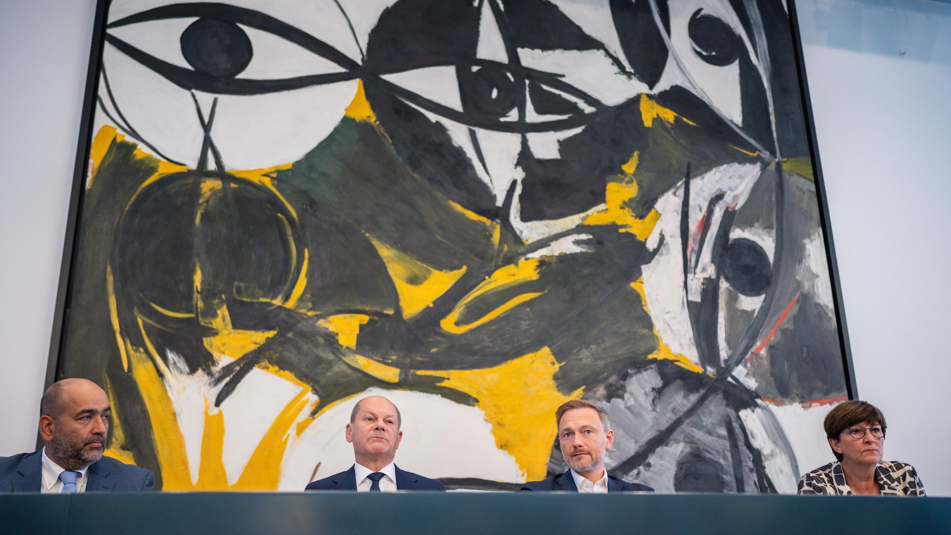 Die vier Politiker sitzen vor einem großen Kunstwerk, das von großen Augen geprägt ist.