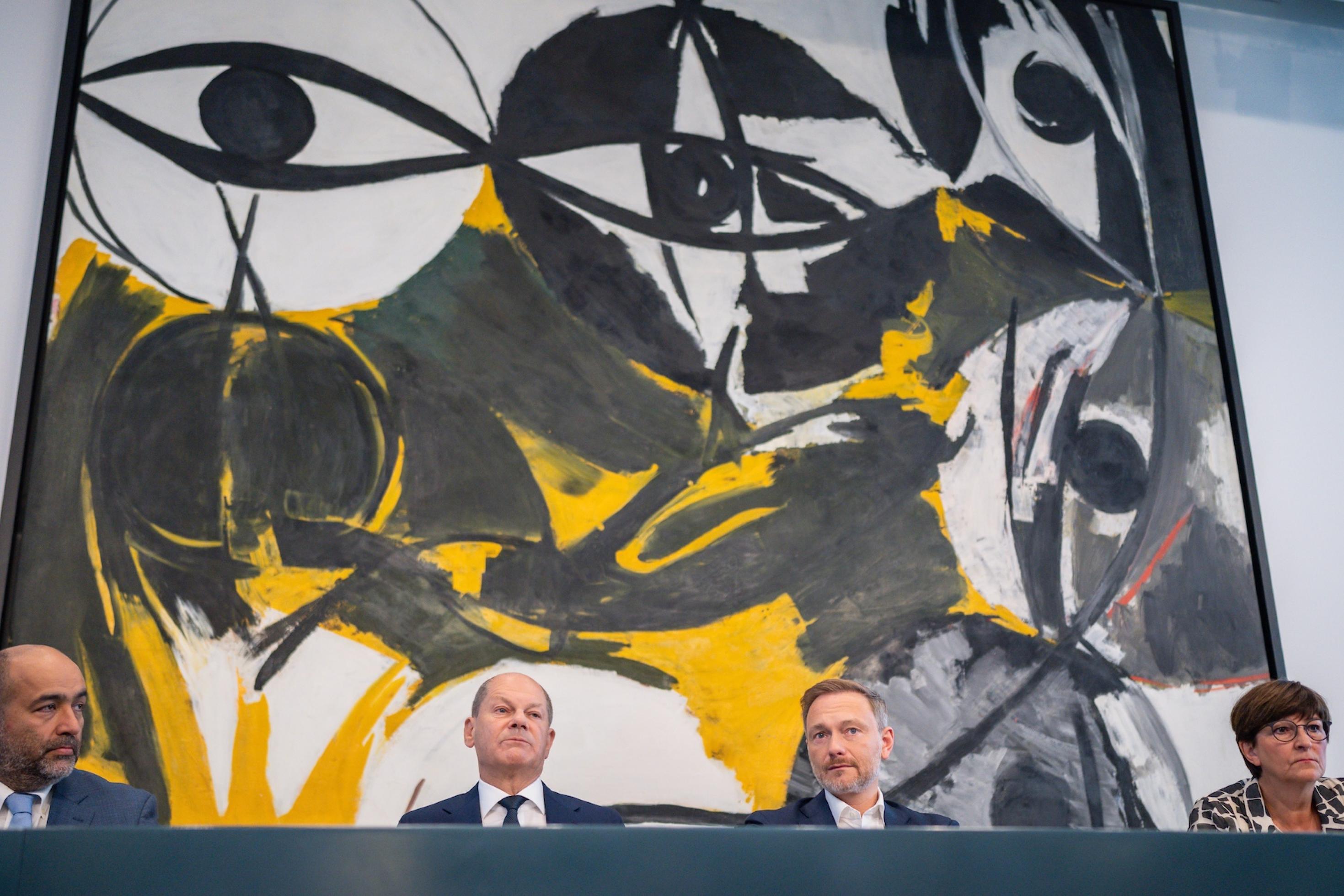 Die vier Politiker sitzen vor einem großen Kunstwerk, das von großen Augen geprägt ist.