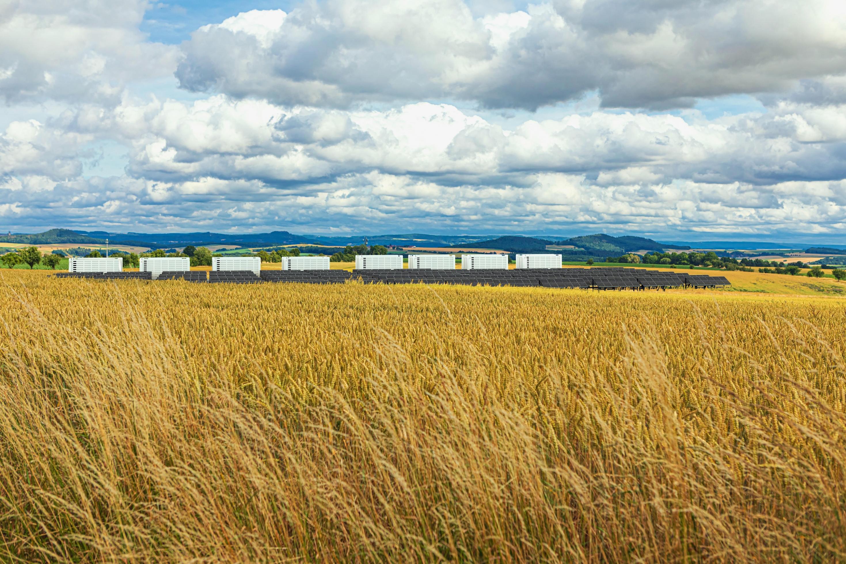 Eine weiter Landschaft mit Getreidefeld und Mittelgebirge, darin ein Solarpark mit Großbatterien als weißen Containern